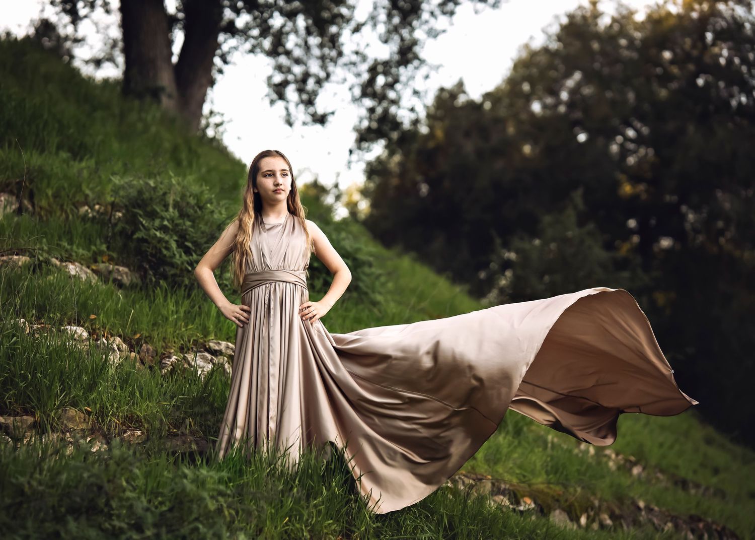 A flowing beige dress billows dramatically in the wind on a grassy hillside during golden hour.