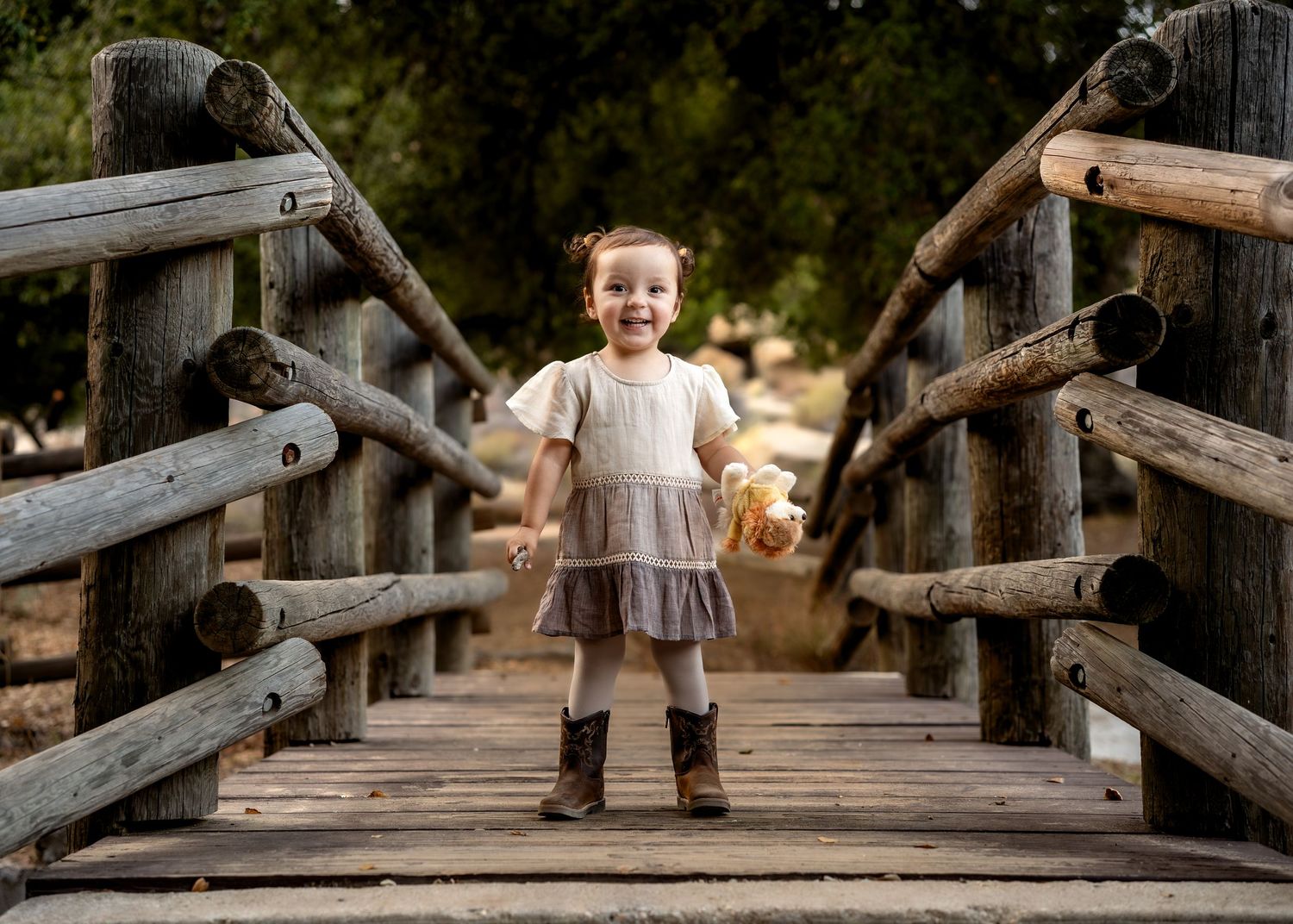A small child stands smiling on a rustic wooden footbridge in a natural outdoor setting.