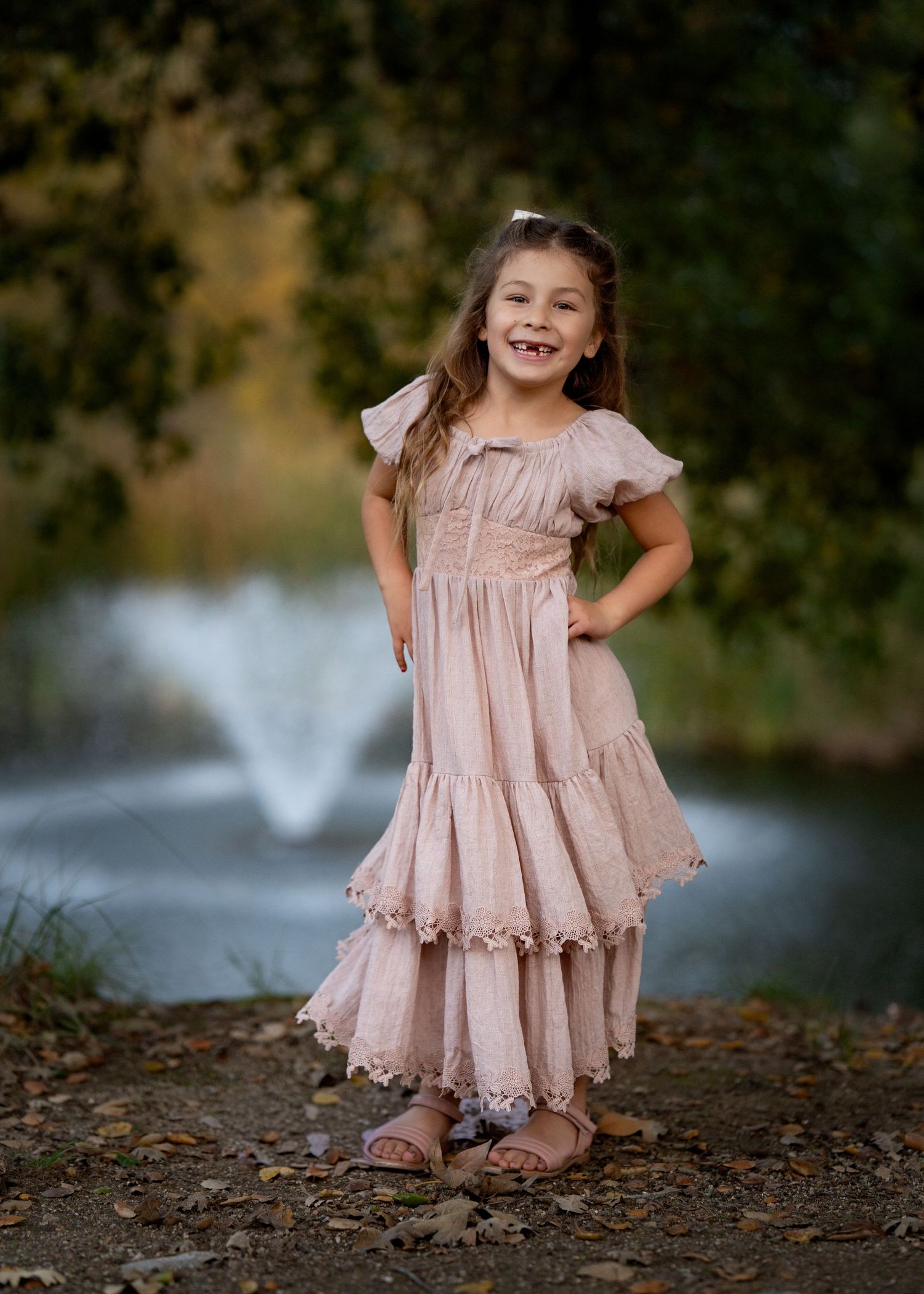 Child in pink lace dress twirling and playing near water fountain.
