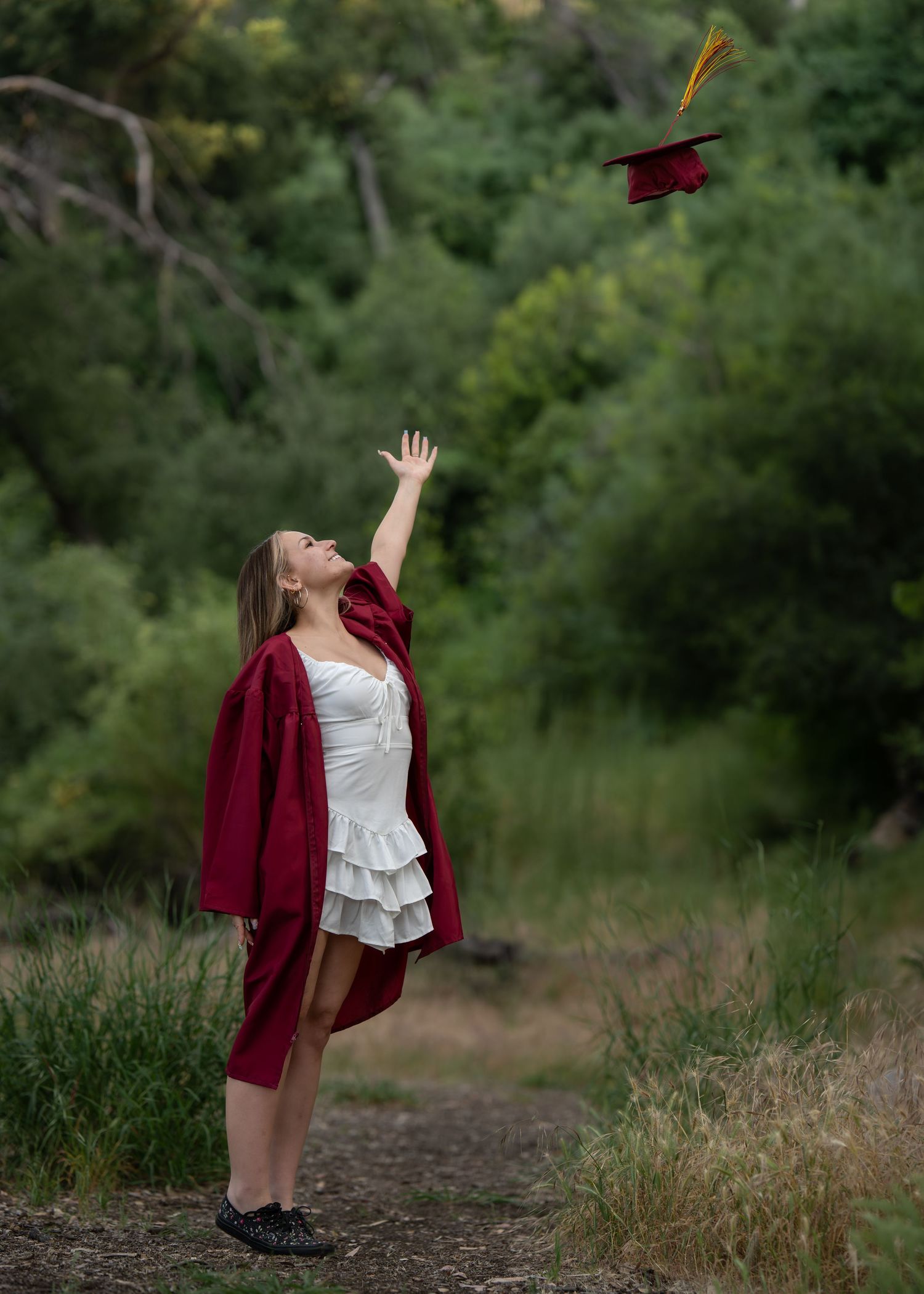 A young woman in a red graduation gown tosses her cap into the air outdoors, celebrating her achievement.