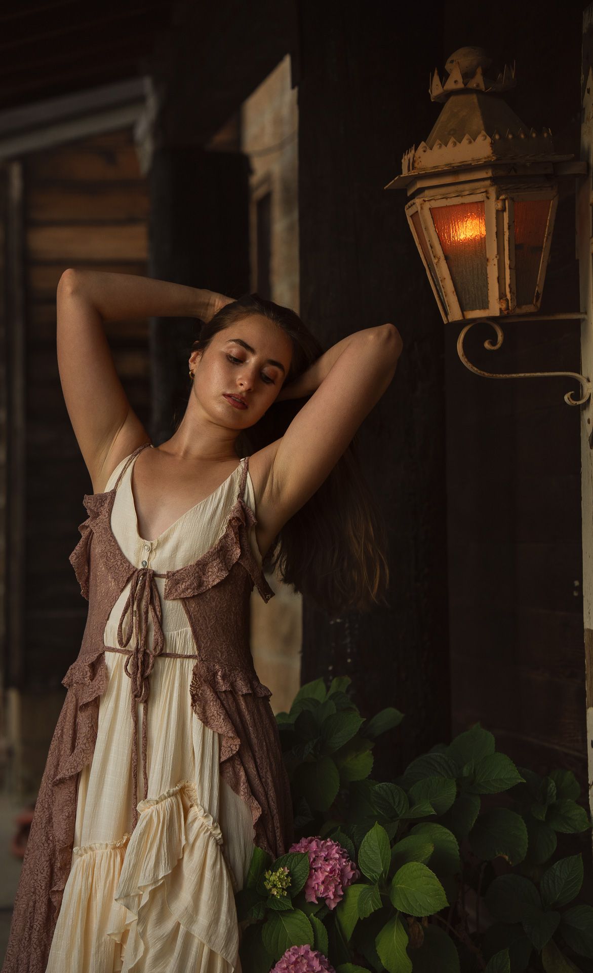 Young woman in vintage cream and brown ruffled dress poses by a glowing antique lantern near rustic wooden building.