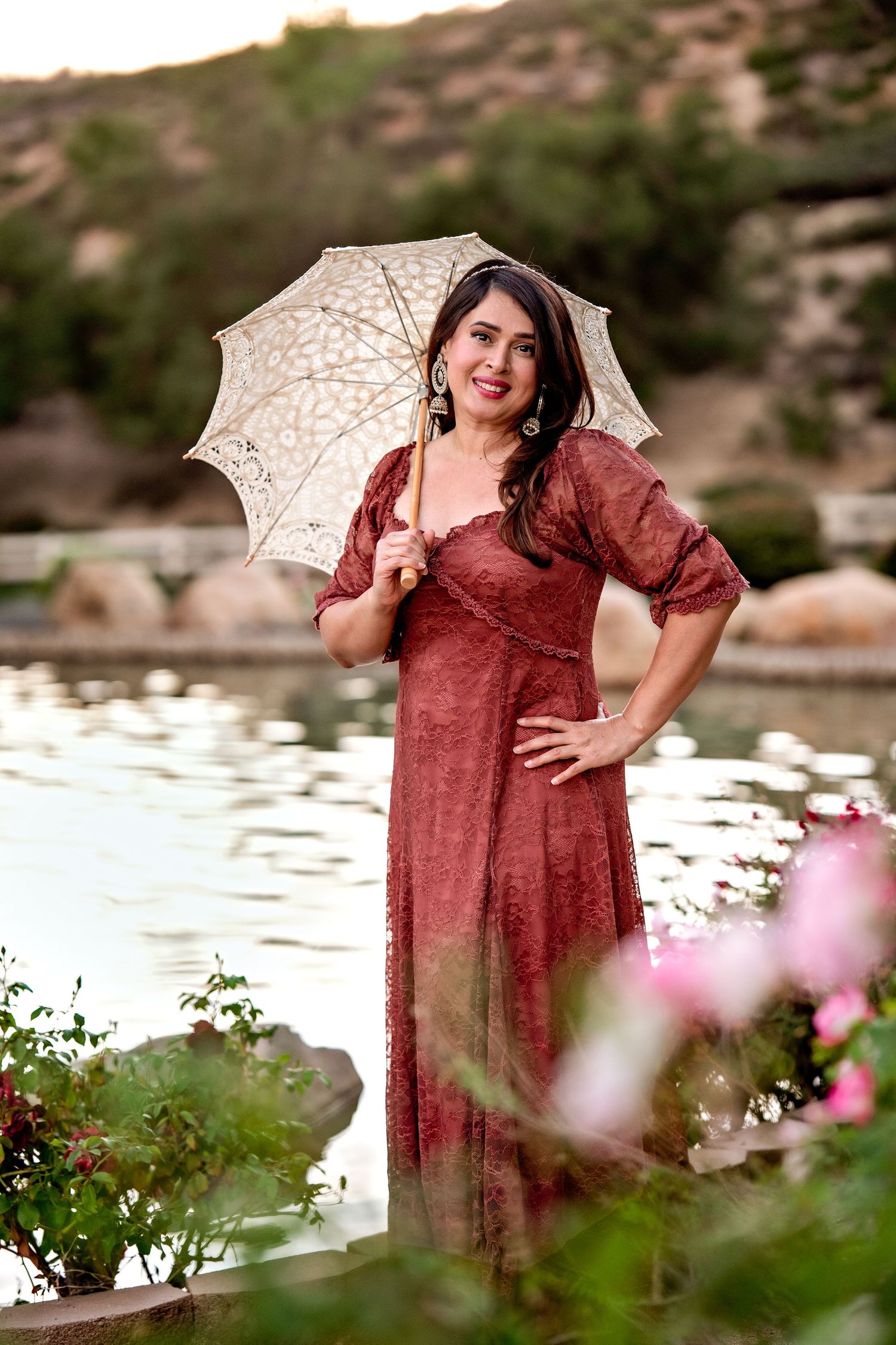 A person in a burgundy dress poses with a white parasol by a serene lake surrounded by blooming flowers.