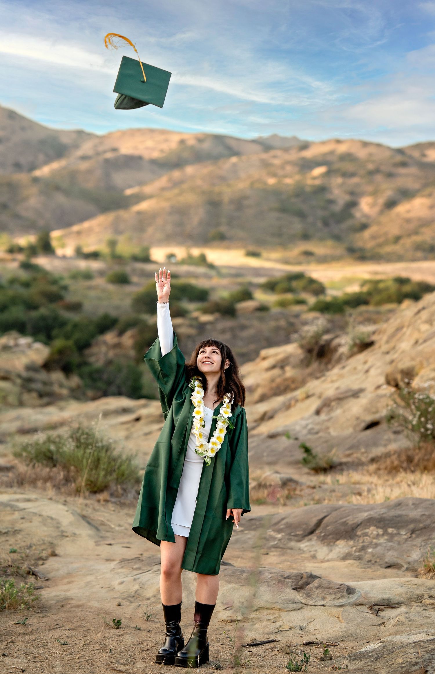 Graduate in green cap and gown poses against mountainous landscape.