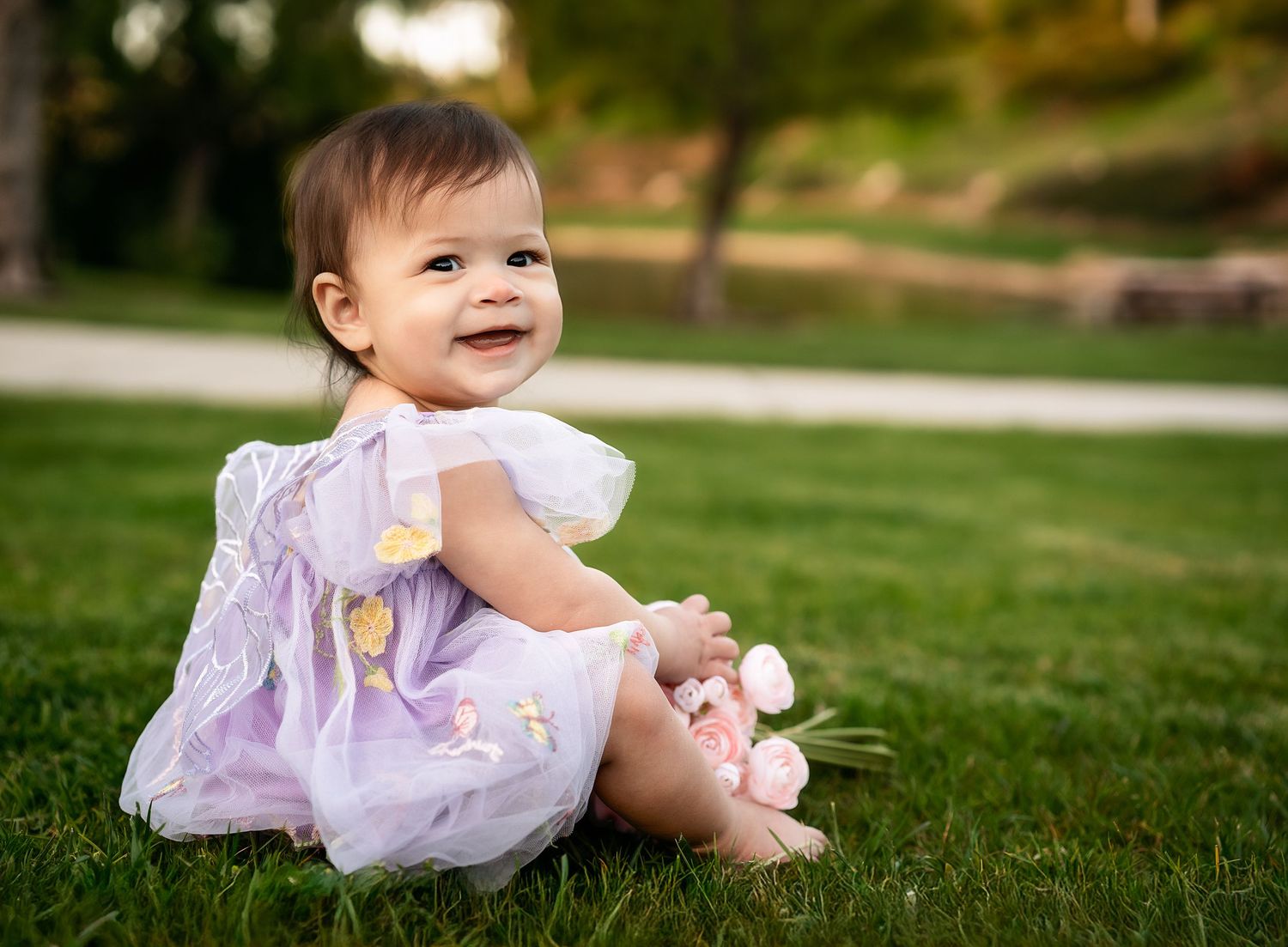 Baby in purple dress sitting on grass with a sweet smile.