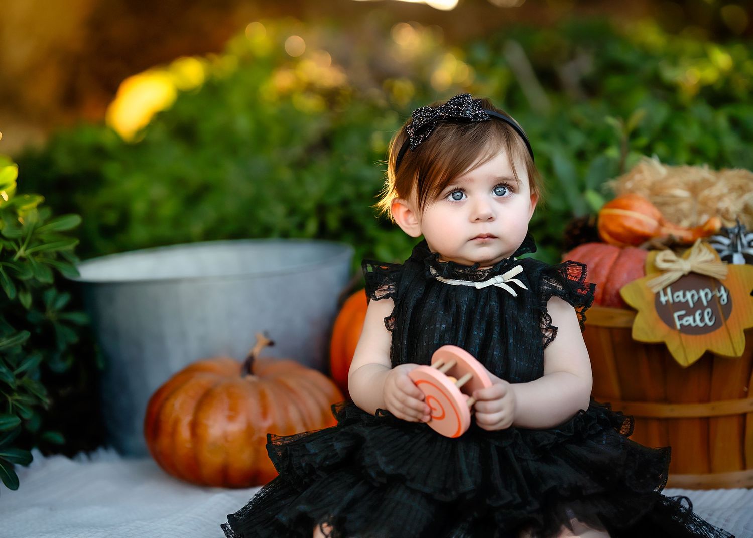 Baby in black dress sitting among fall decorations and pumpkins outdoors.