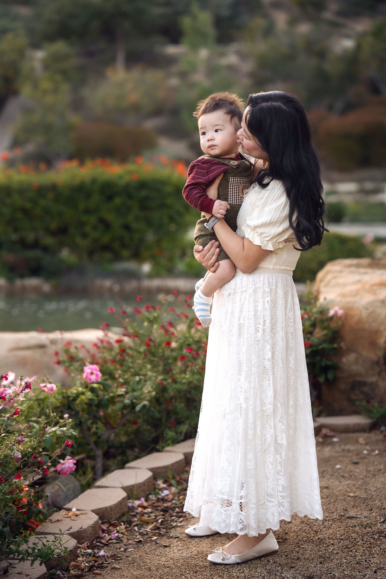 A woman in a white lace dress holds a small child in a garden setting with pink flowers.