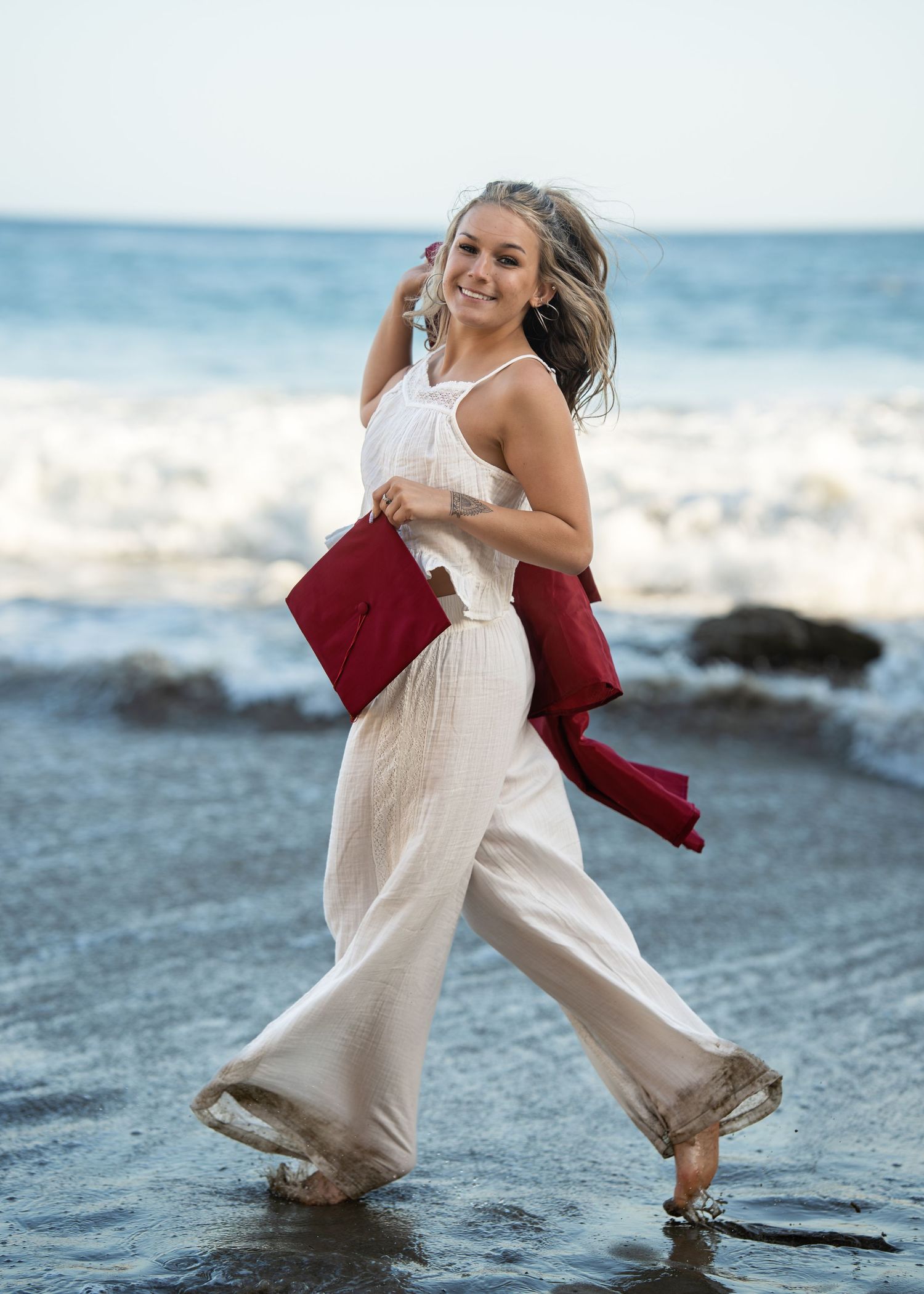 Smiling woman in white jumpsuit holding red jacket walks barefoot on a beach with ocean waves behind her.