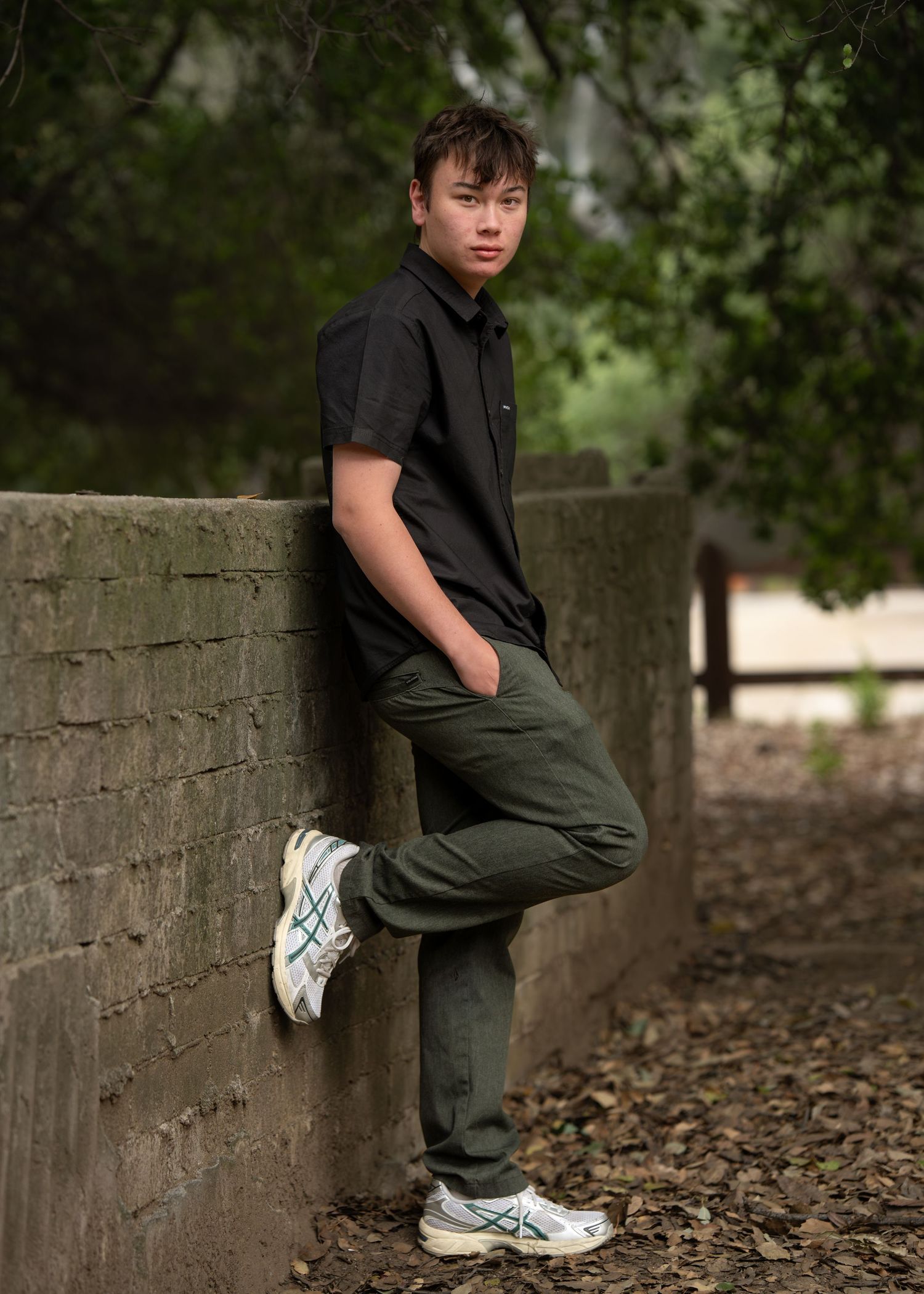 Young man in black shirt and olive pants leaning against stone wall outdoors wearing ASICS sneakers.