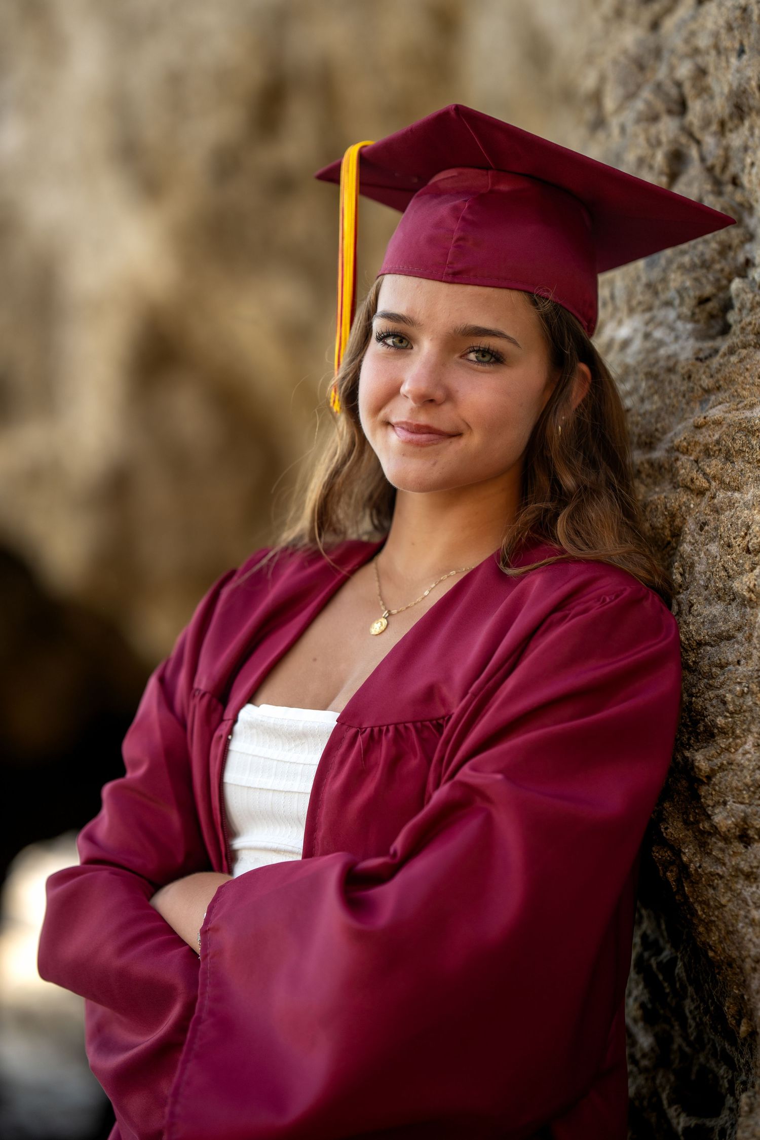 Young woman in maroon graduation cap and gown smiles with arms crossed, leaning against a stone wall outdoors.