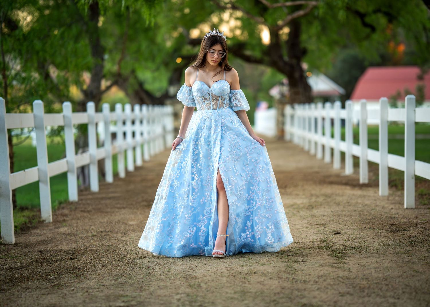 Woman in light blue off-shoulder lace ball gown with slit poses on a tree-lined path by a white fence.