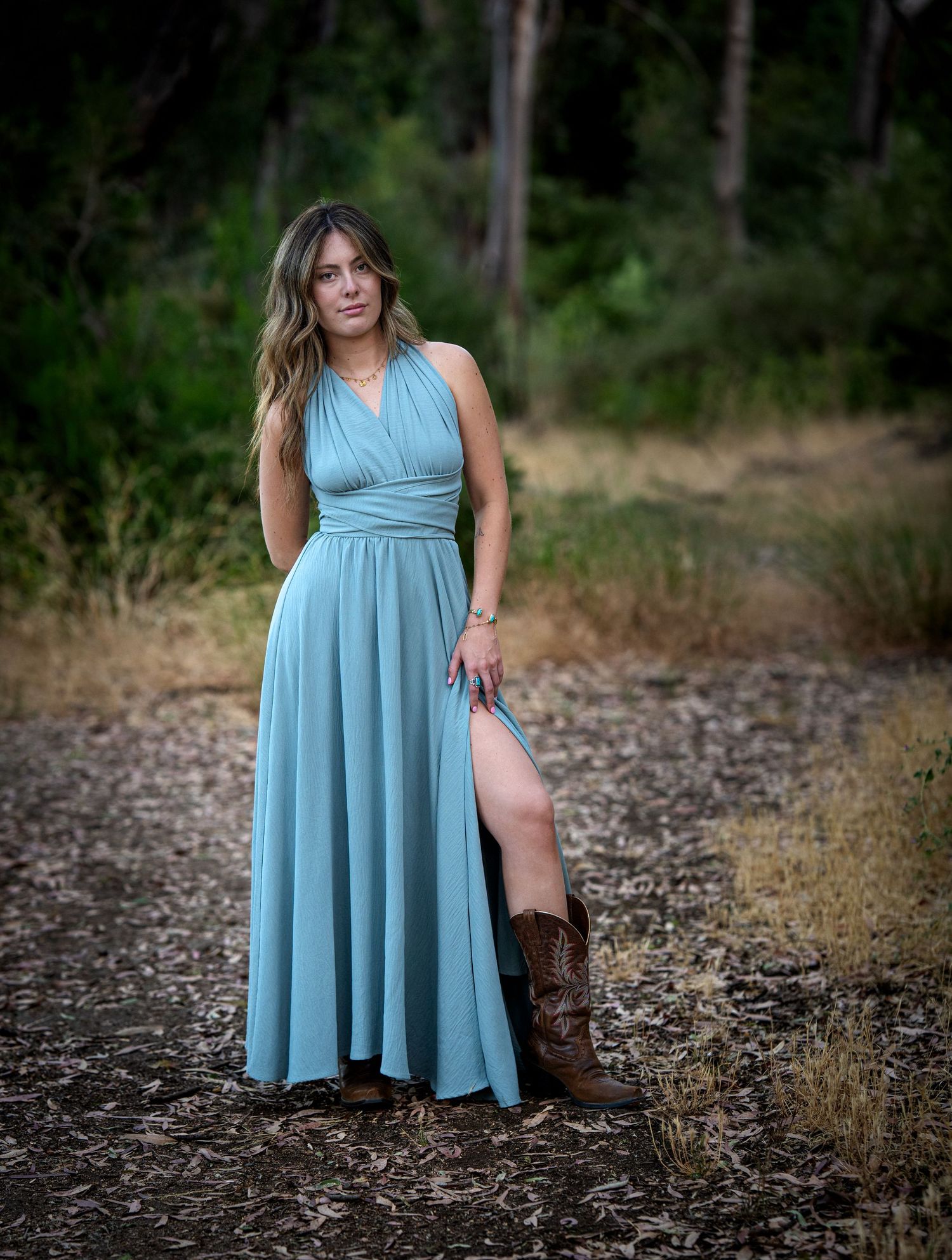 Woman in teal maxi dress and cowboy boots poses on a forest path for a portrait photo.