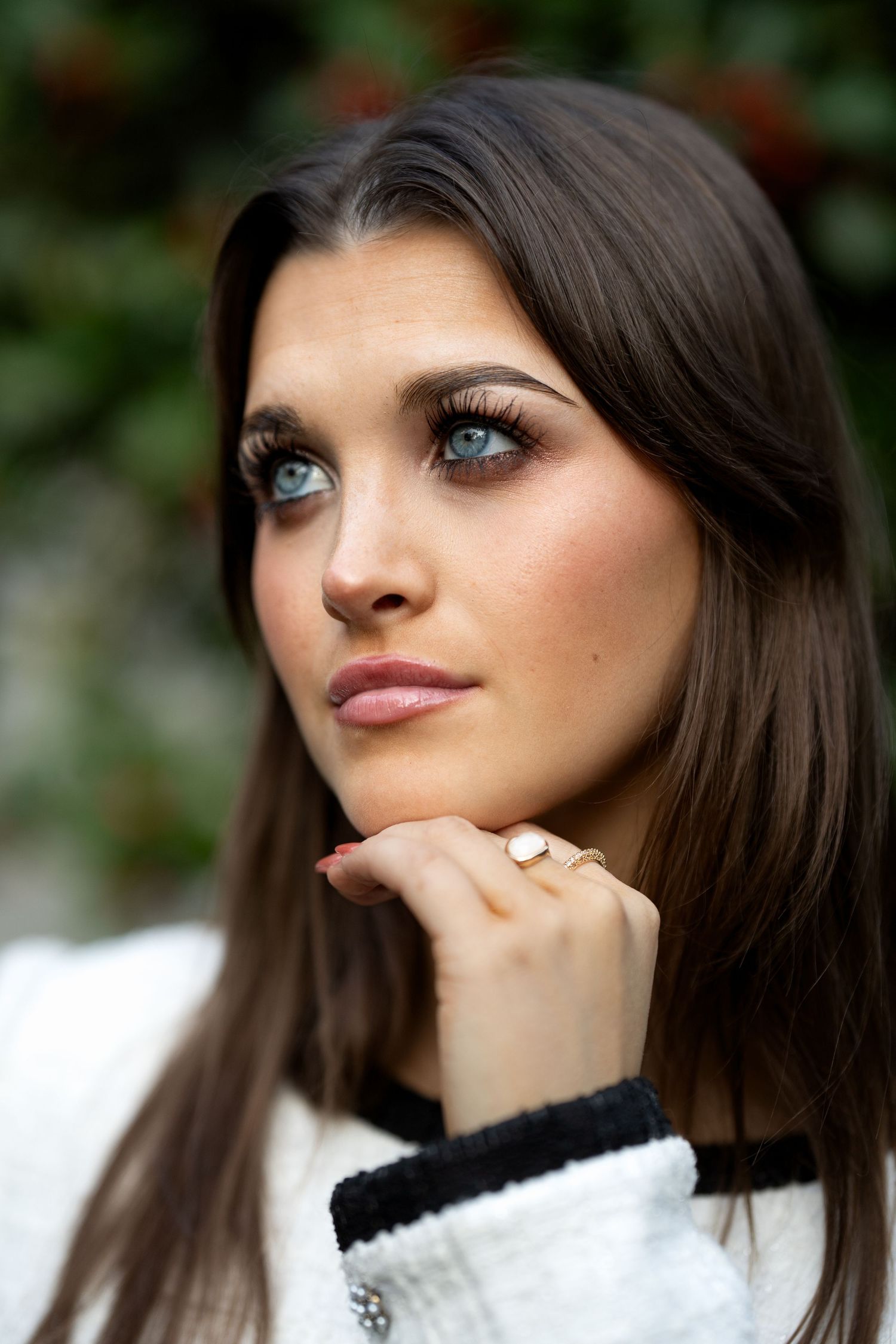 Young woman with blue eyes and dark hair resting chin on hand, wearing a white jacket and gold ring outdoors.