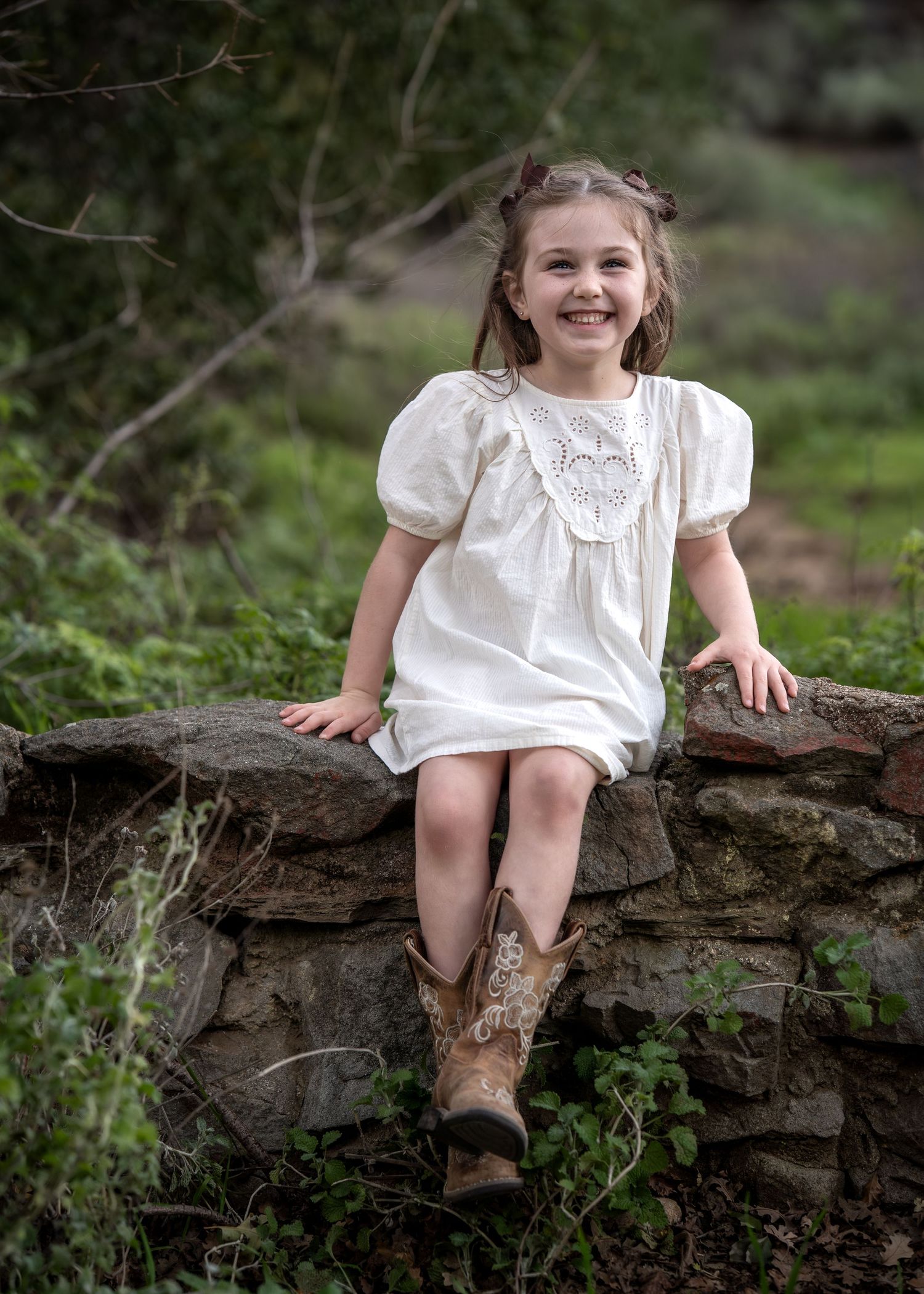 A child in a white dress sits casually on a rustic log in a wooded setting.