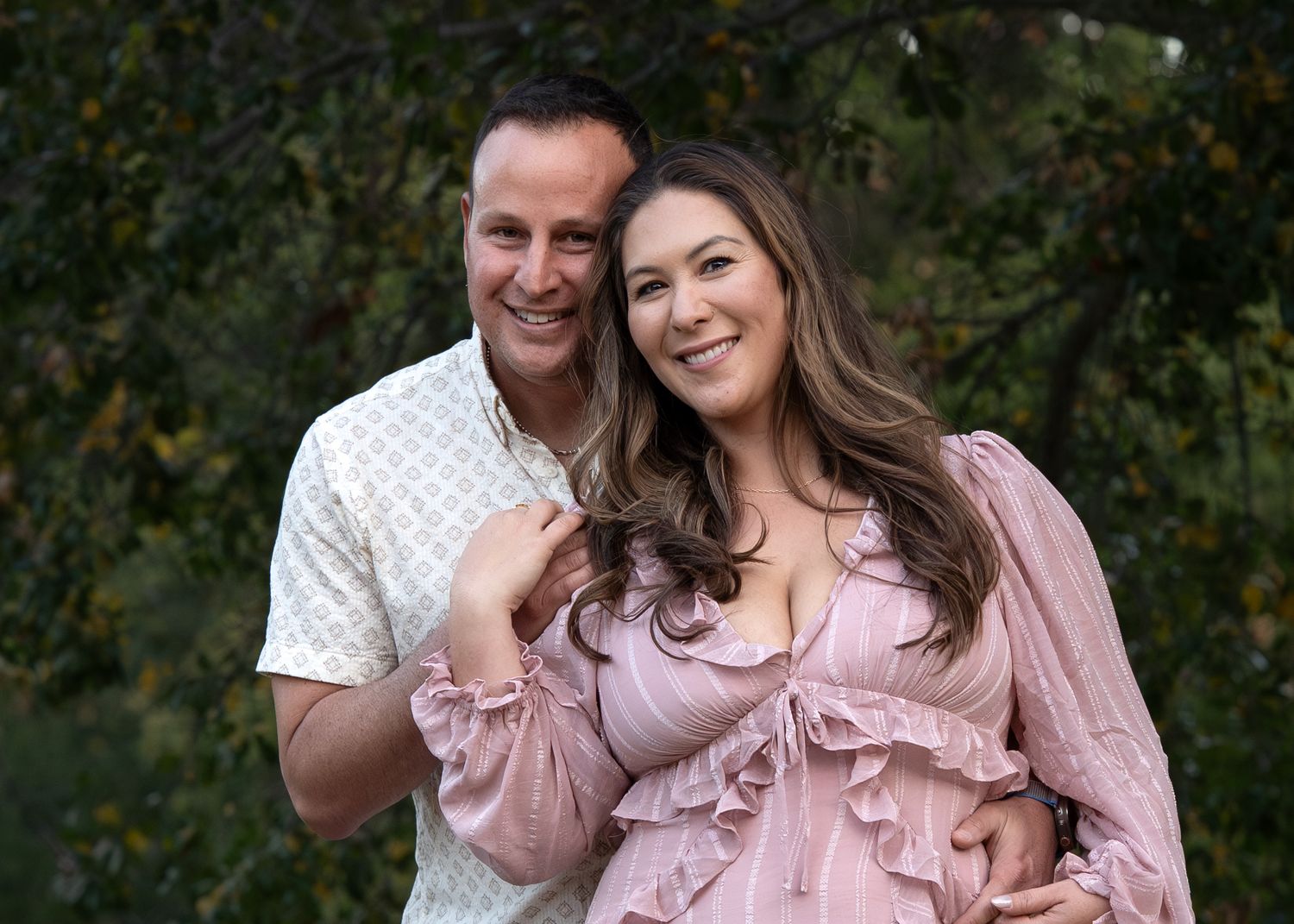 Happy couple in outdoor portrait with woman wearing pink ruffled dress against natural backdrop.