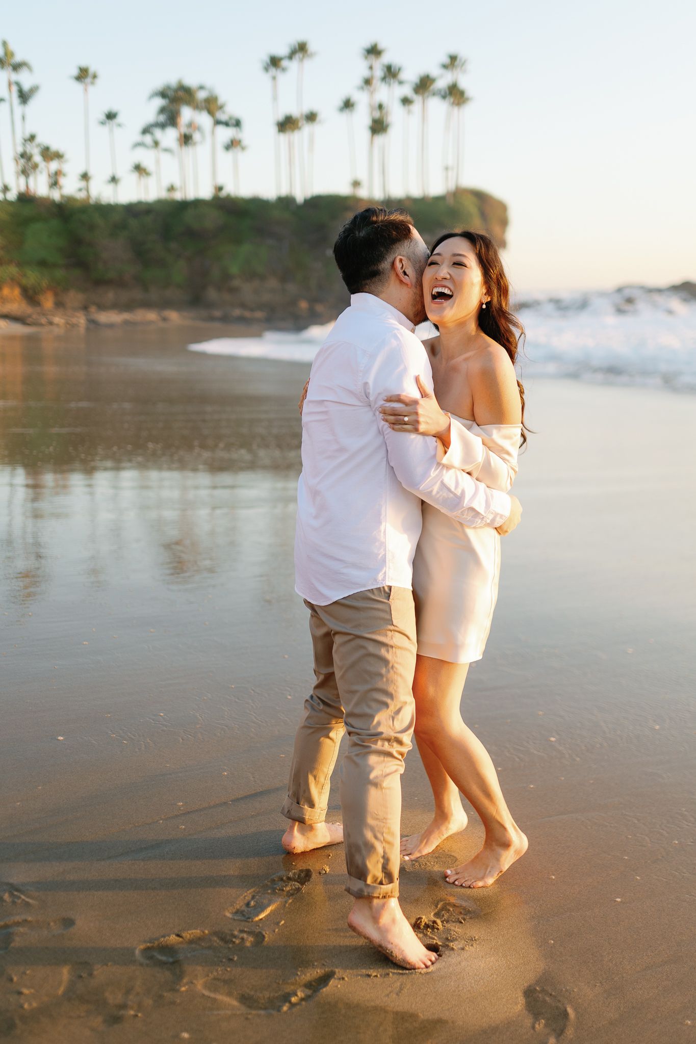 A laughing couple embraces on a wet sandy beach at golden hour with palm trees visible in the background.
