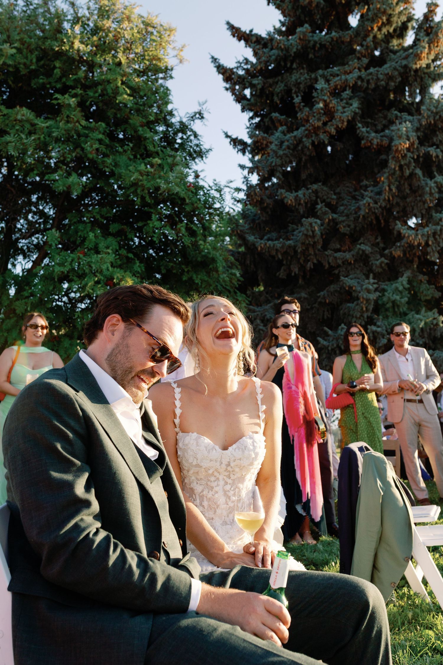 Bride laughs joyfully beside groom in green suit during outdoor wedding reception on grass.