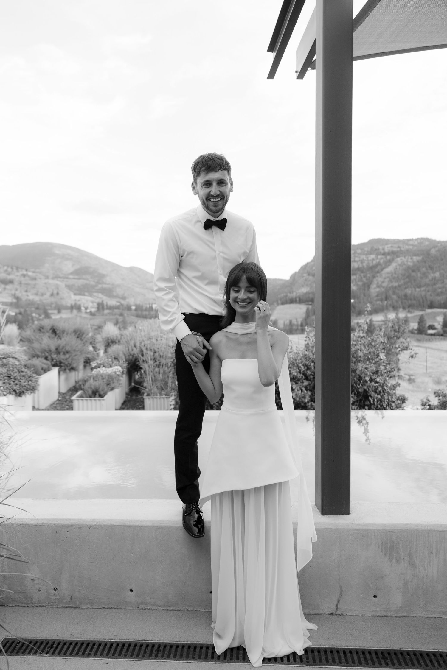 Smiling bride and groom posing on an outdoor terrace with scenic mountain views in black and white wedding photo.