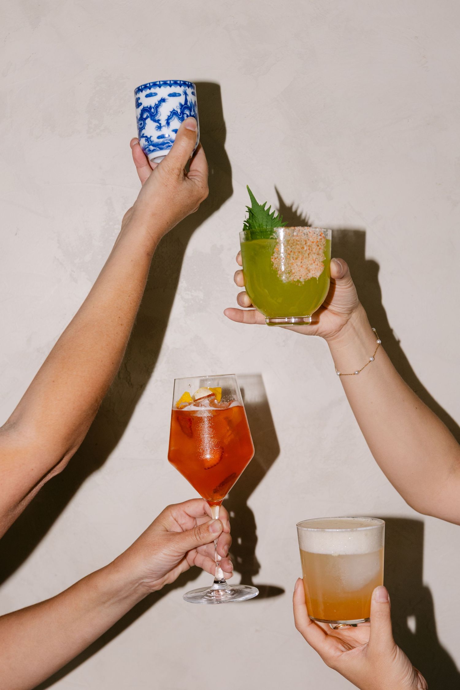 Four hands holding colorful cocktails including a wine glass, ceramic cup, and highball glasses against a neutral wall.