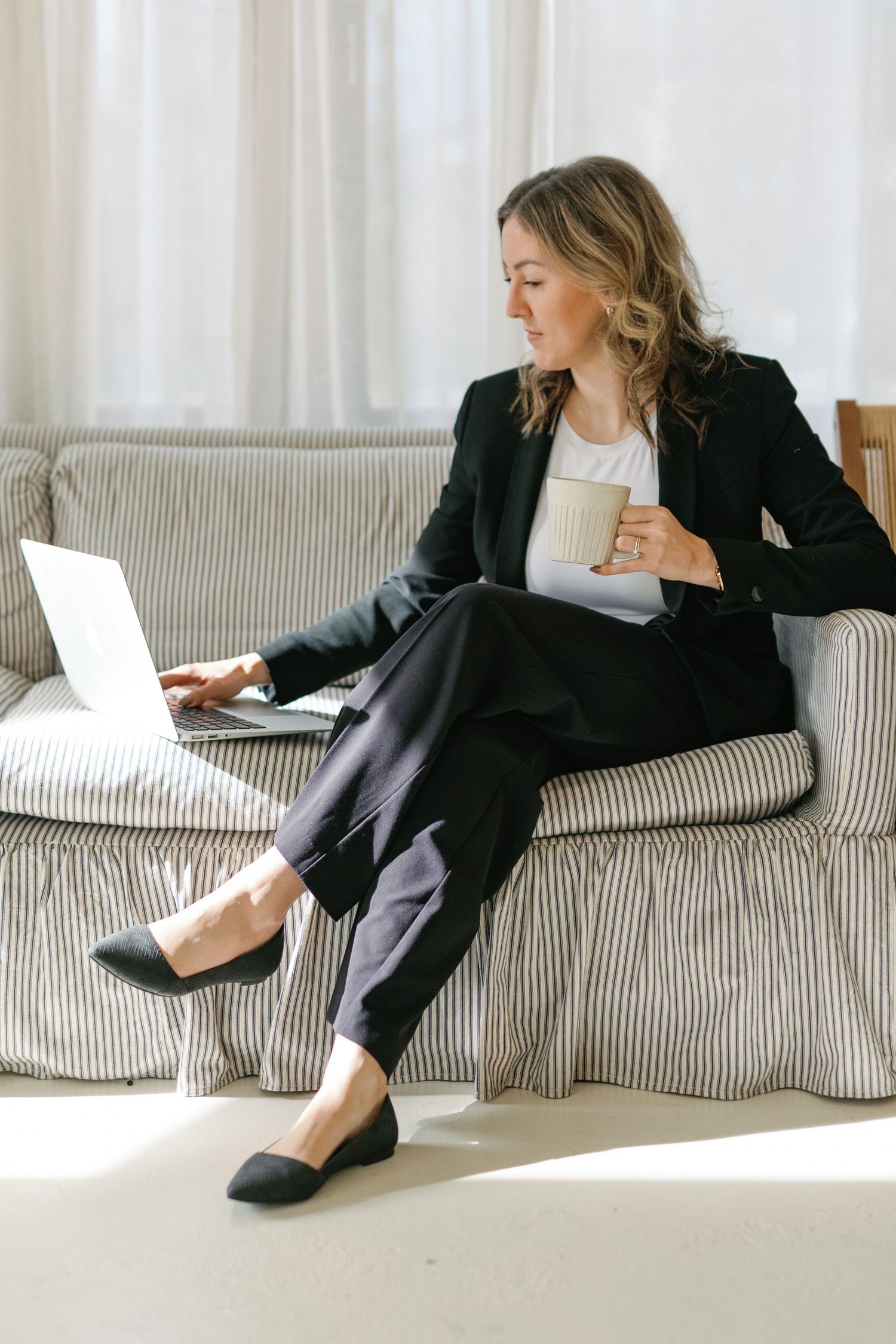Professional woman in black suit working on laptop while holding coffee cup, sitting on striped sofa.