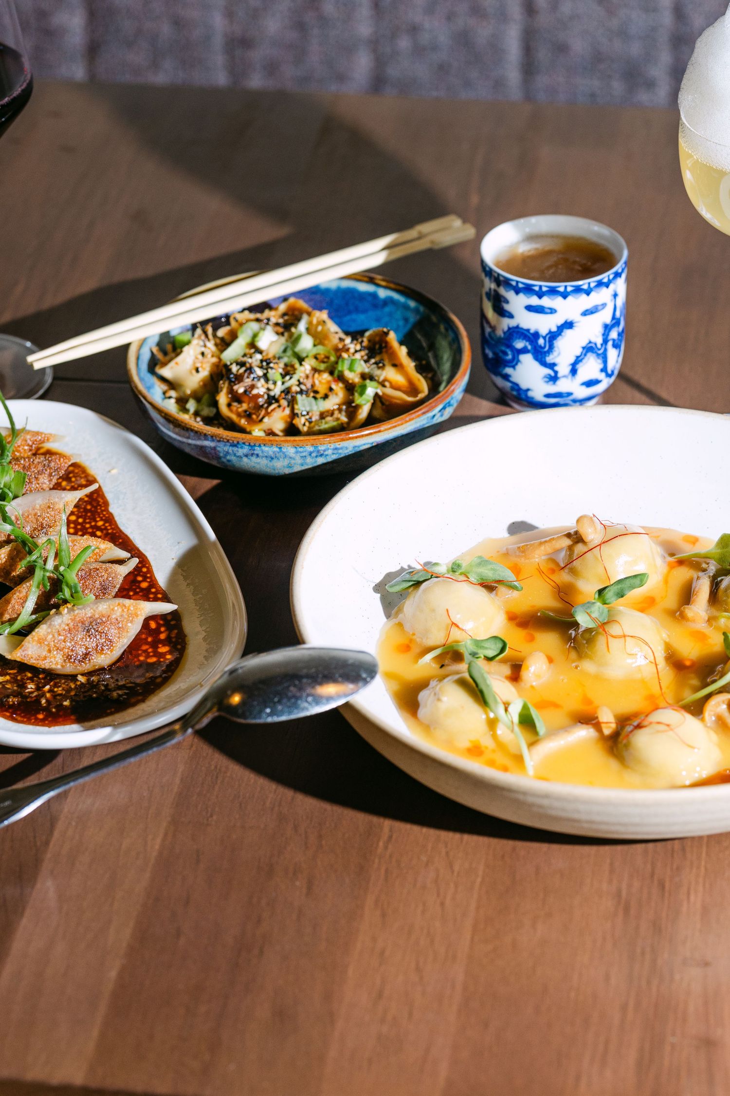 Asian dumplings spread across three dishes with chili oil, orange sauce, and sesame, served with chopsticks and green tea.