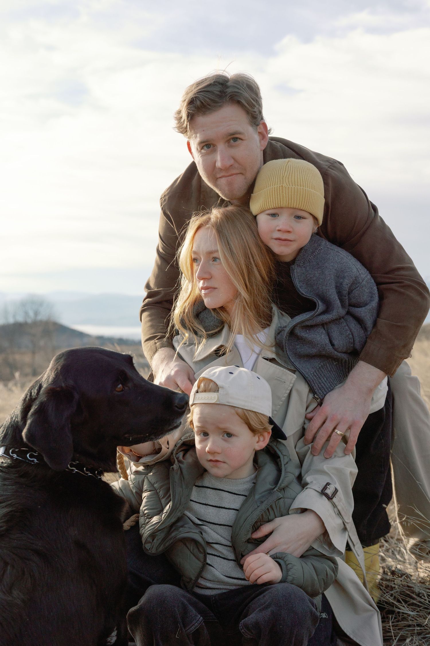 Family of four with black Labrador dog posing outdoors in a field on a cloudy day.
