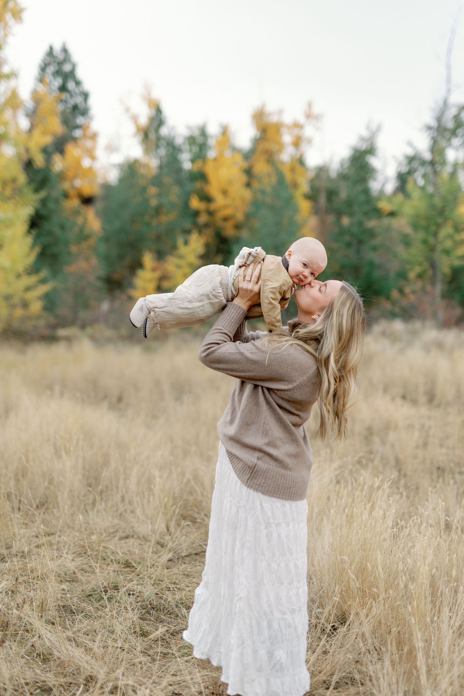 Mother lifting smiling baby in autumn field with colorful trees, wearing neutral tones in a warm fall outdoor portrait.
