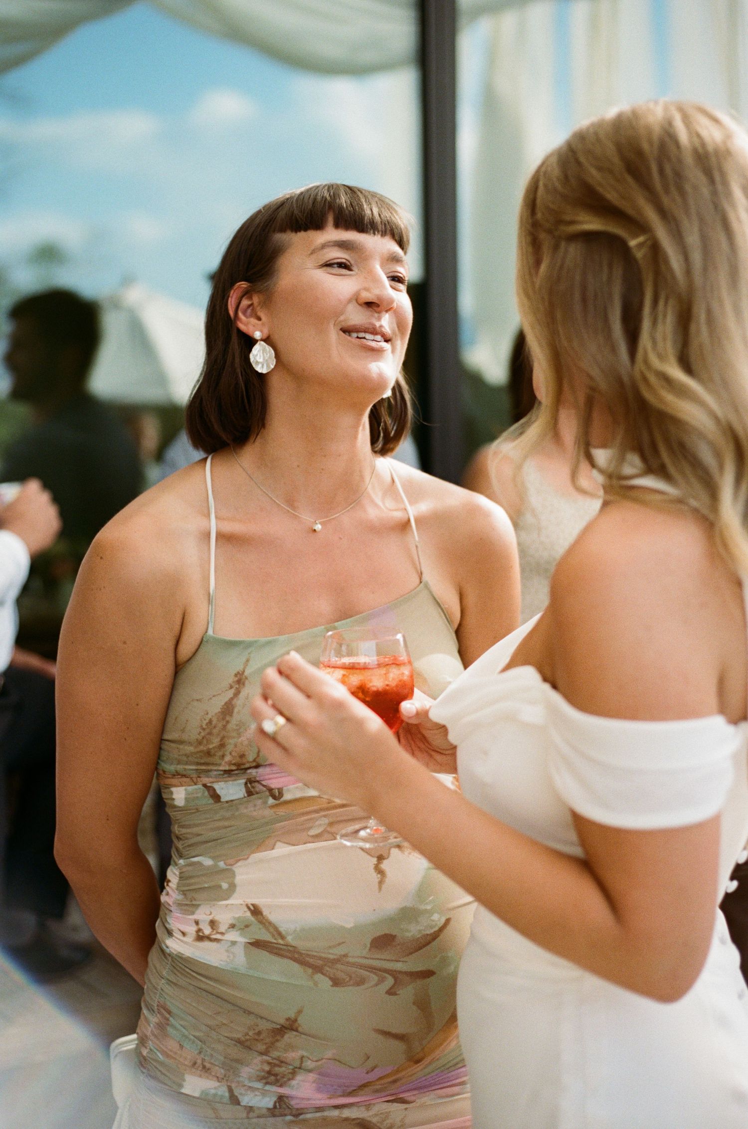 A smiling wedding guest in a floral dress chats with the bride during a sunny outdoor reception.