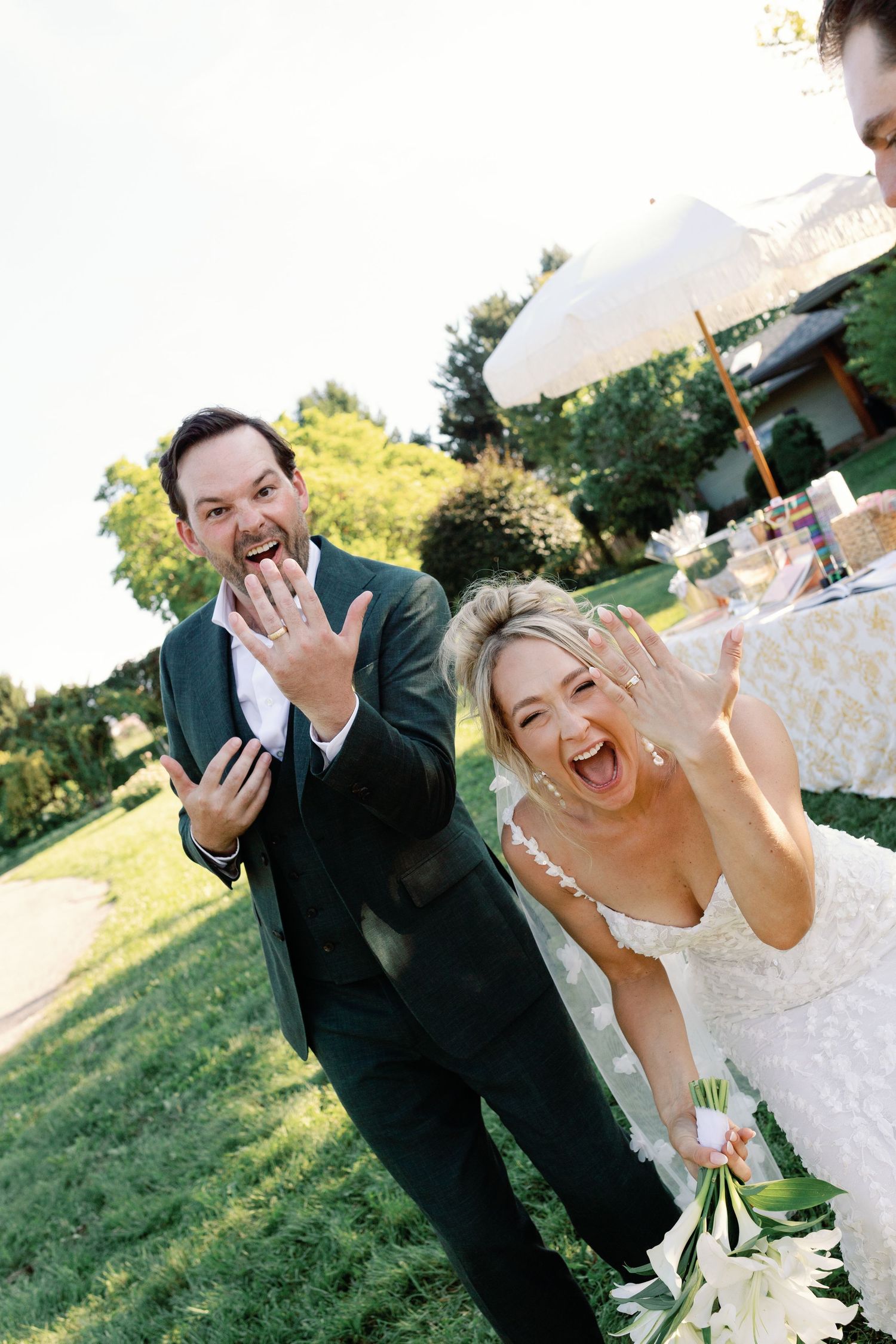 Joyful newlyweds show off wedding rings laughing at outdoor garden ceremony.