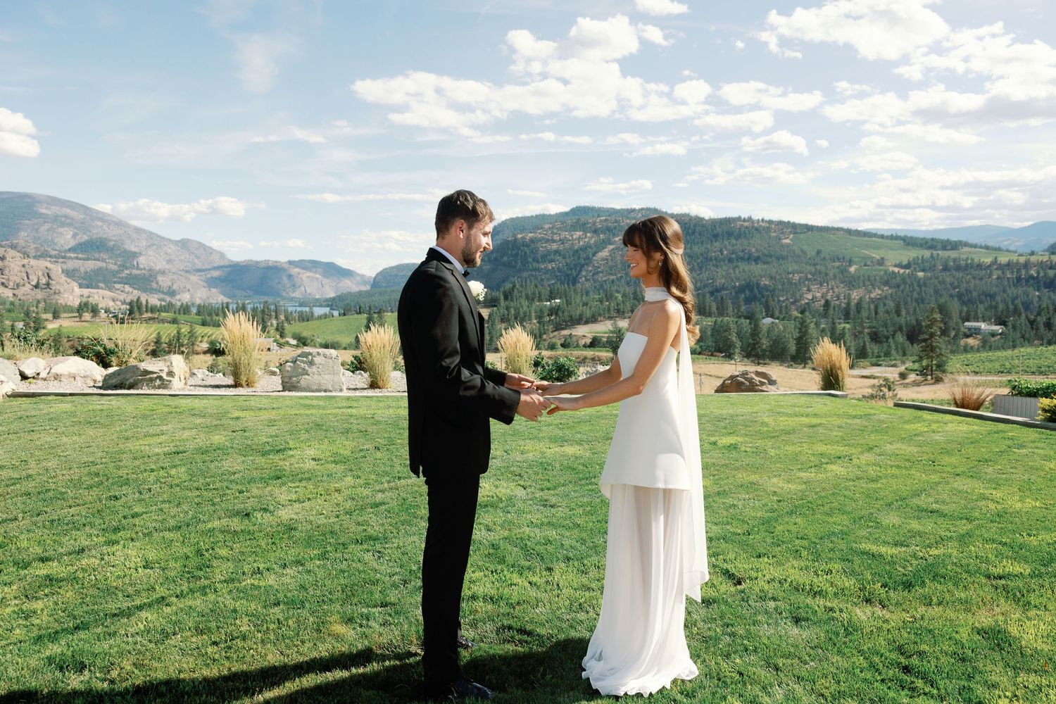 Bride and groom hold hands during outdoor wedding ceremony on lush green lawn with mountains.