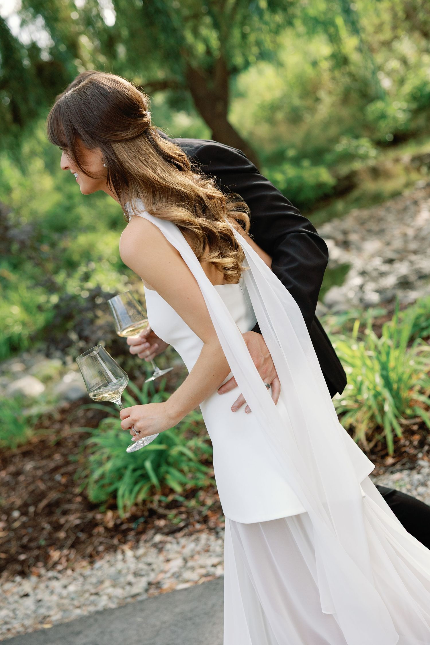Groom playfully dips bride holding champagne glass while strolling through lush green garden at outdoor wedding.