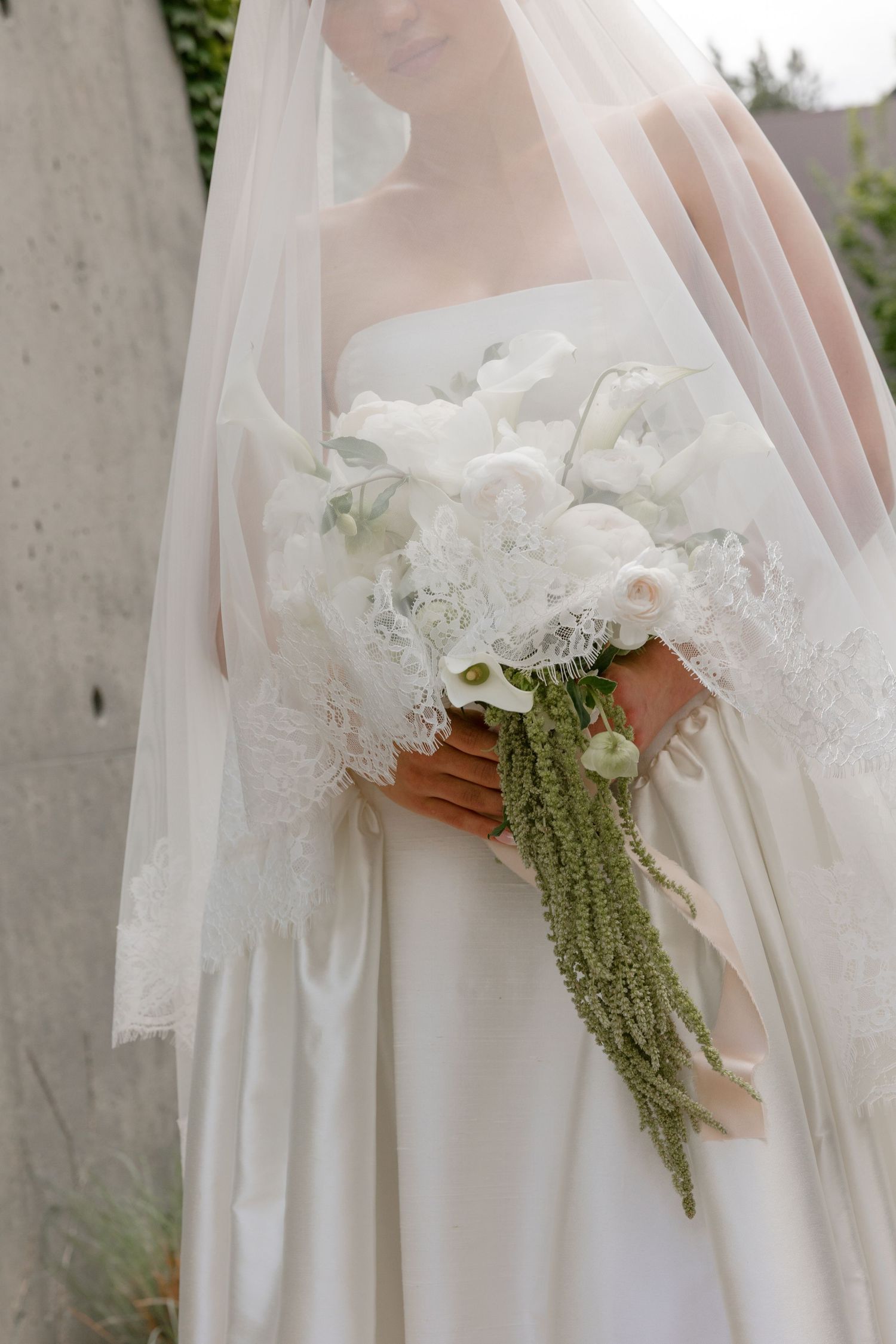 Bride in white gown and lace veil holding elegant bouquet of white roses, calla lilies, and trailing green amaranthus.