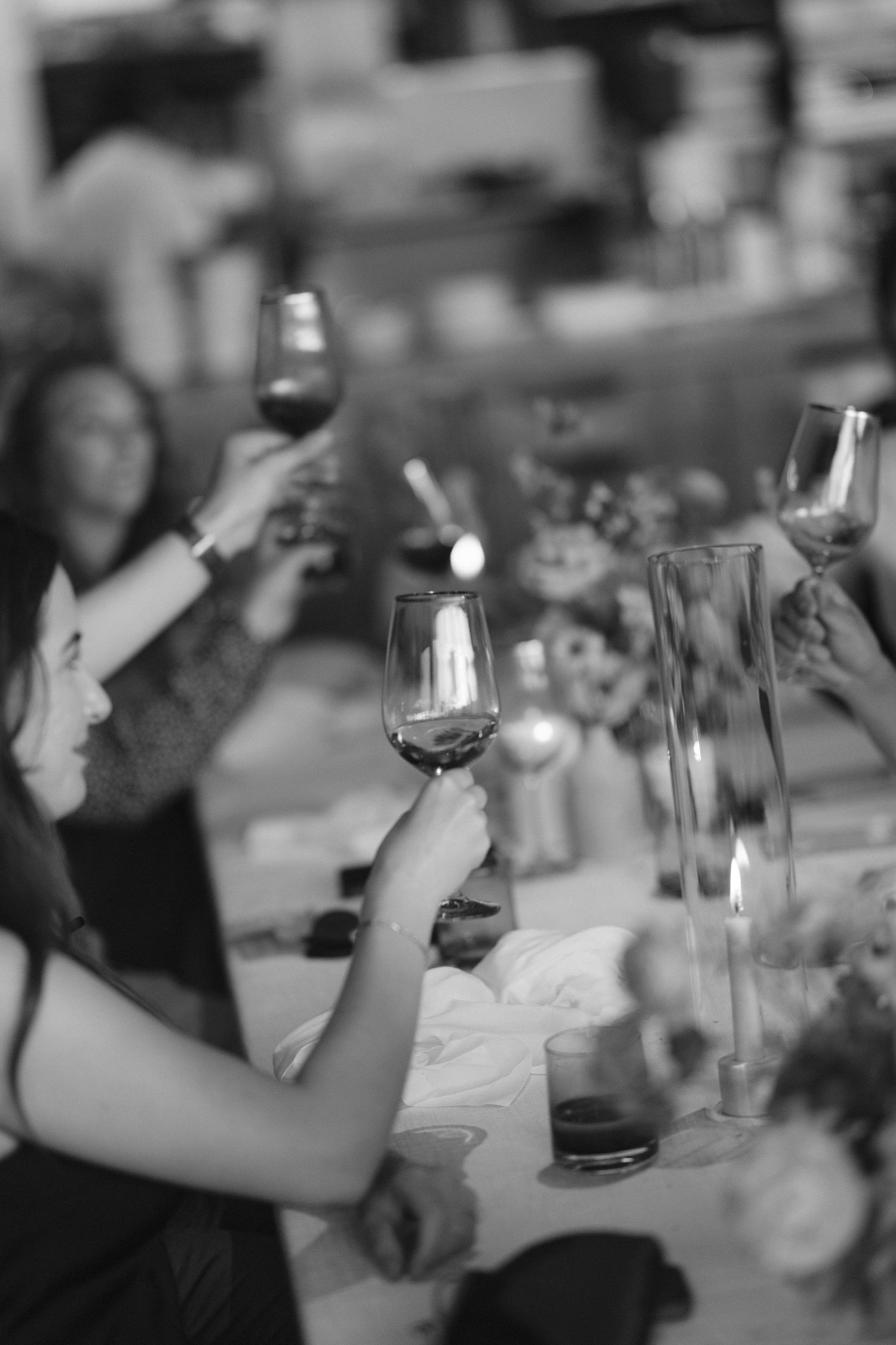 Black and white photo of guests raising wine glasses in a toast at a candlelit dinner party.