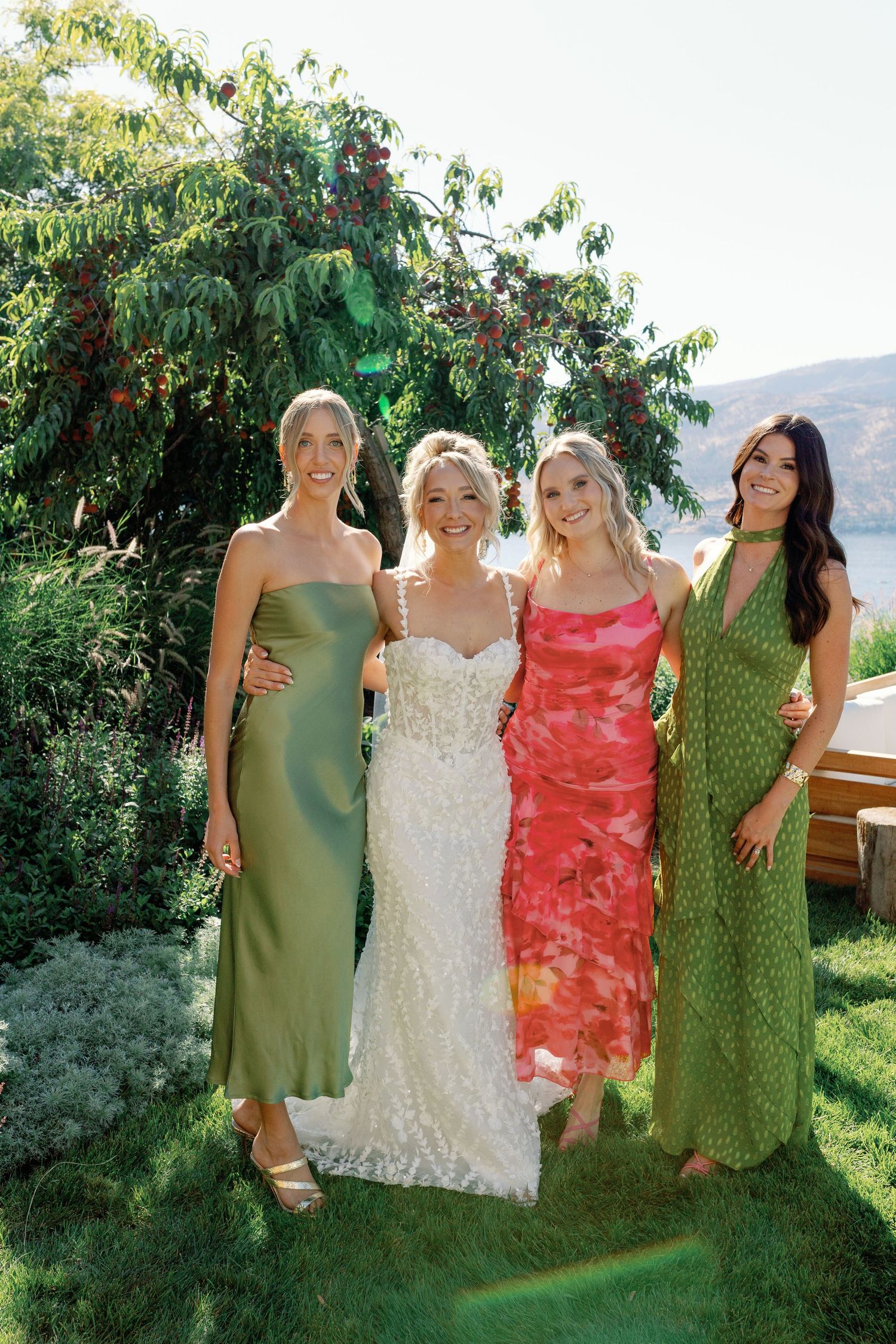 Bride in lace wedding gown poses with three bridesmaids in green and pink dresses in garden setting.