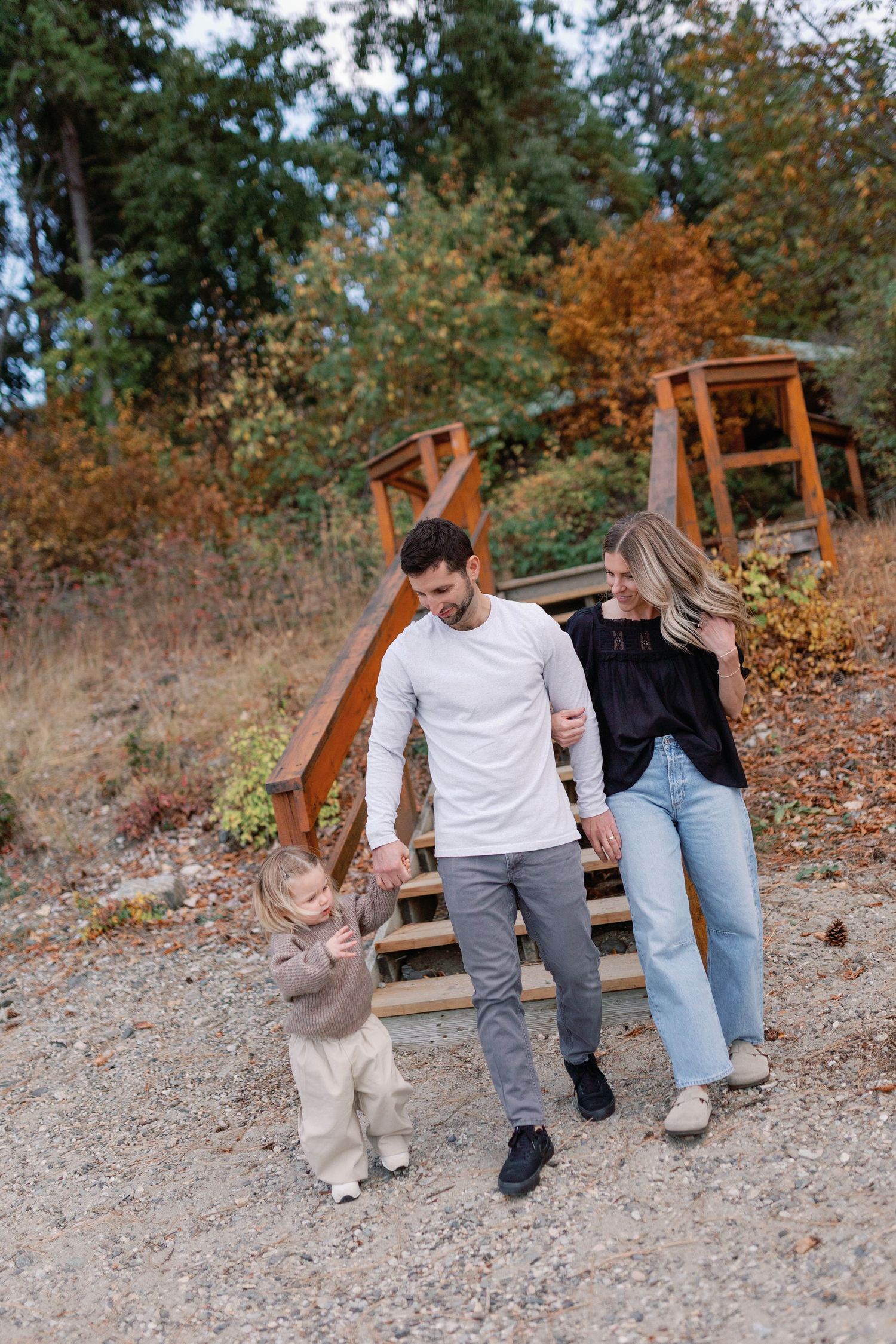 A happy family of three walks outdoors in autumn, parents holding toddler's hands near wooden steps.
