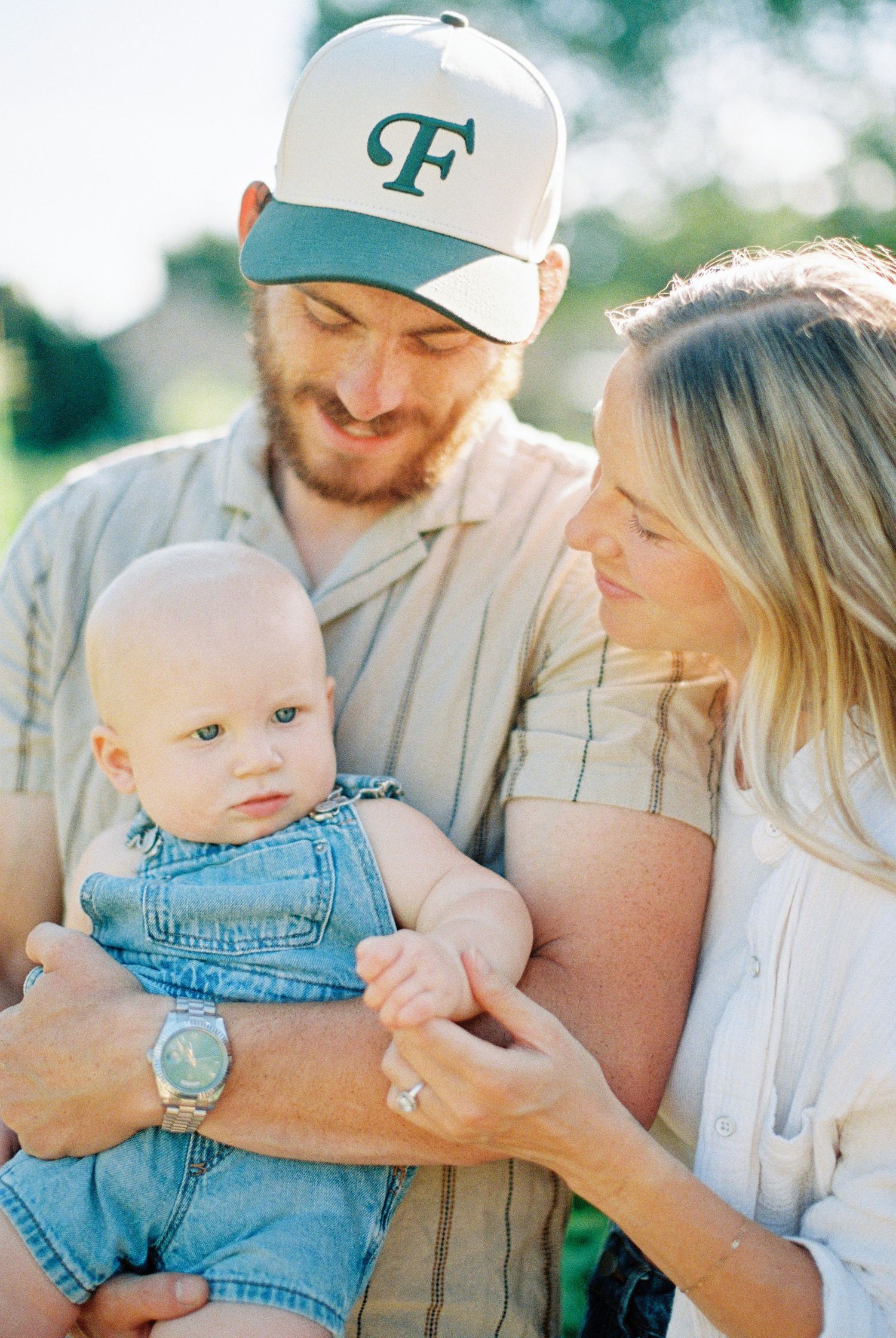 Young parents smiling at baby in denim overalls during golden hour outdoor family portrait.