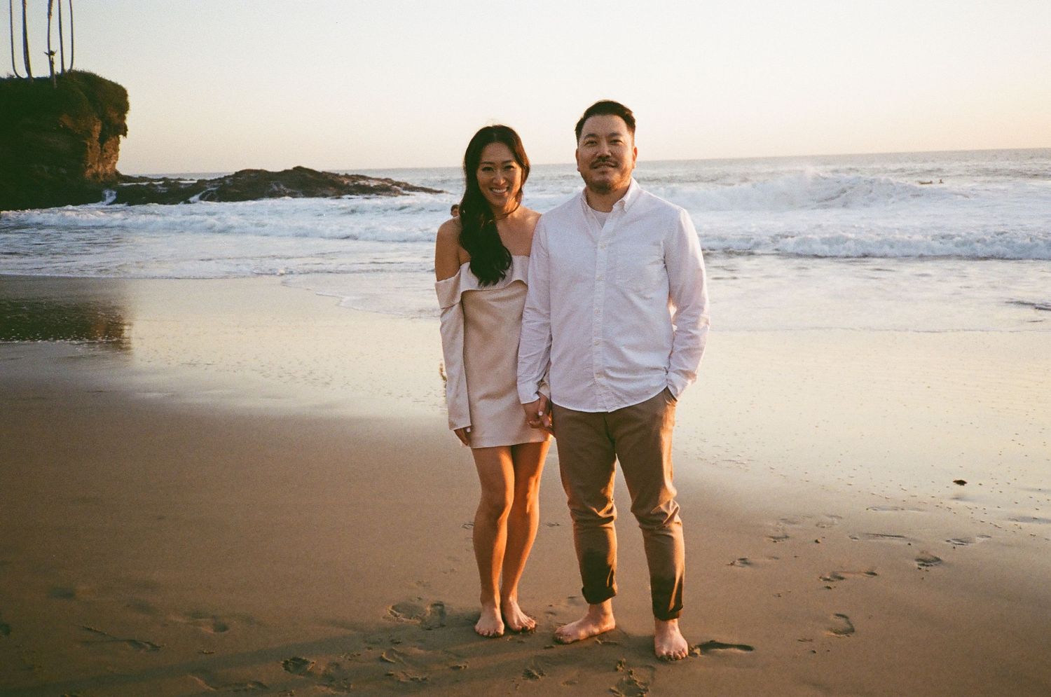 A couple stands together barefoot on a beach at sunset with ocean waves and rocky cliffs in the background.
