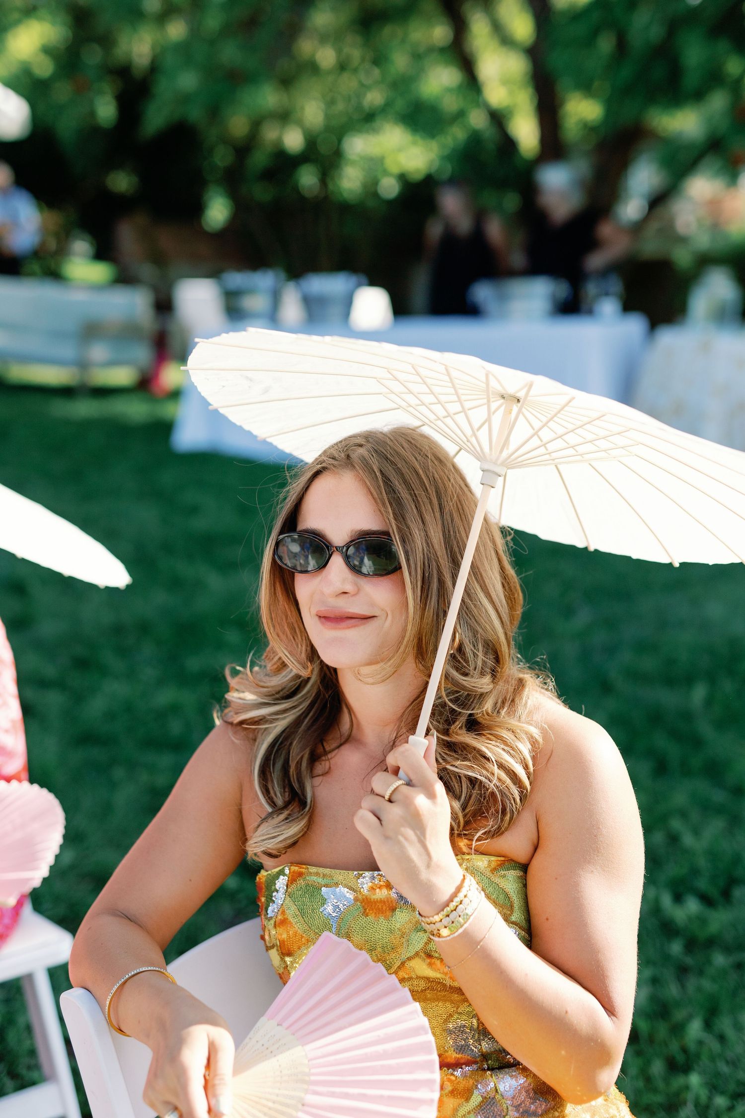 Wedding guest in floral dress holding a parasol and fan at a sunny outdoor ceremony.