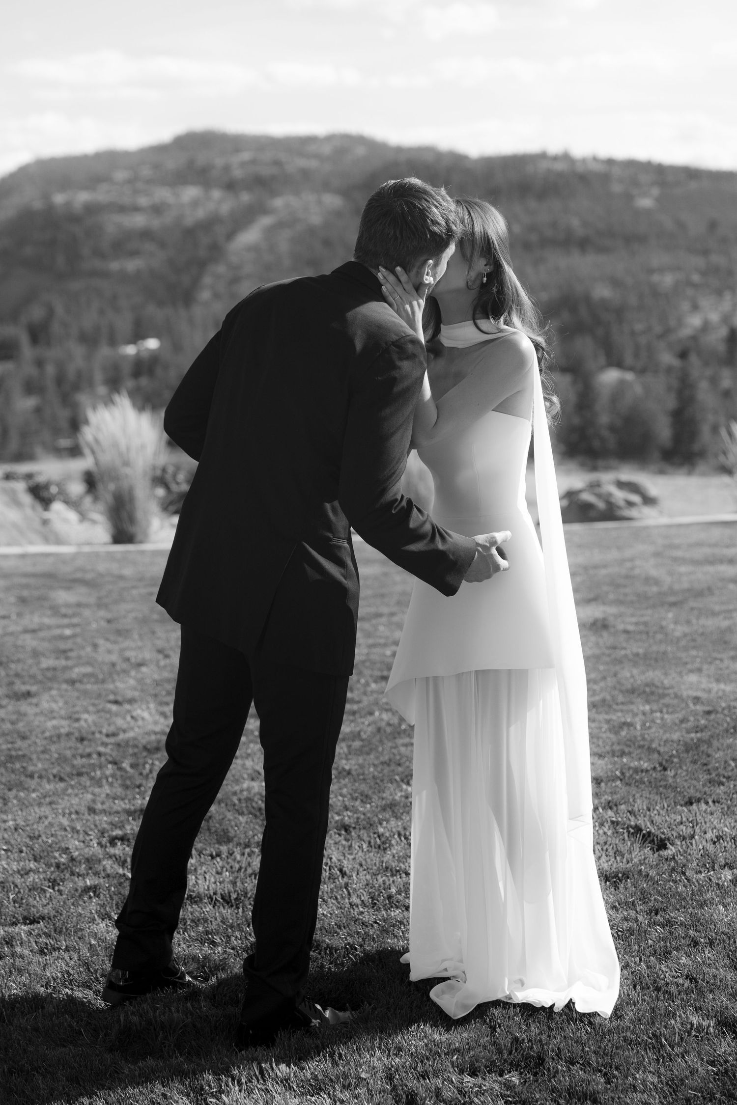 Black and white photo of bride and groom sharing a kiss on green lawn with mountain view.