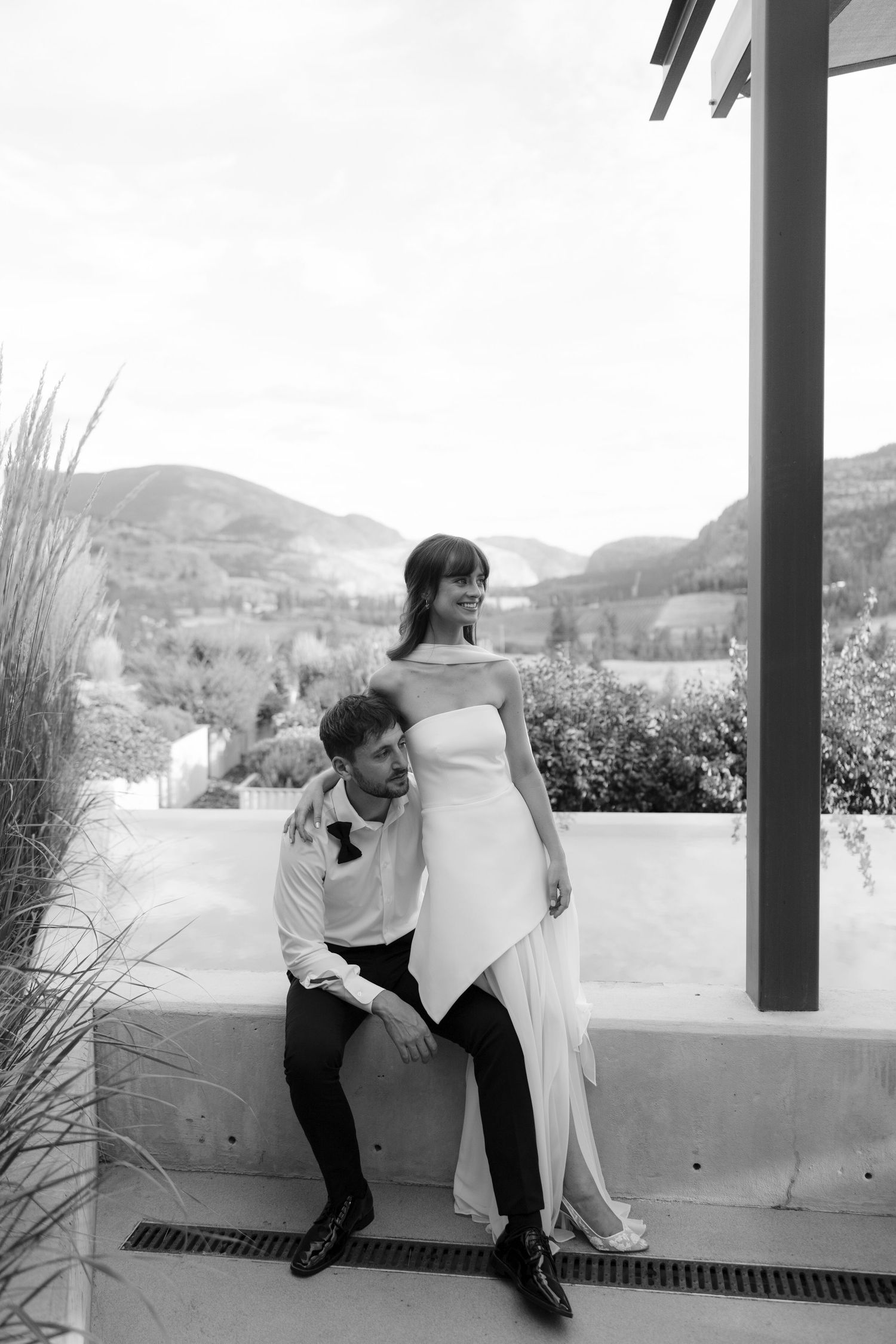 Elegant bride in strapless gown standing beside seated groom in bow tie on outdoor terrace with mountain backdrop.