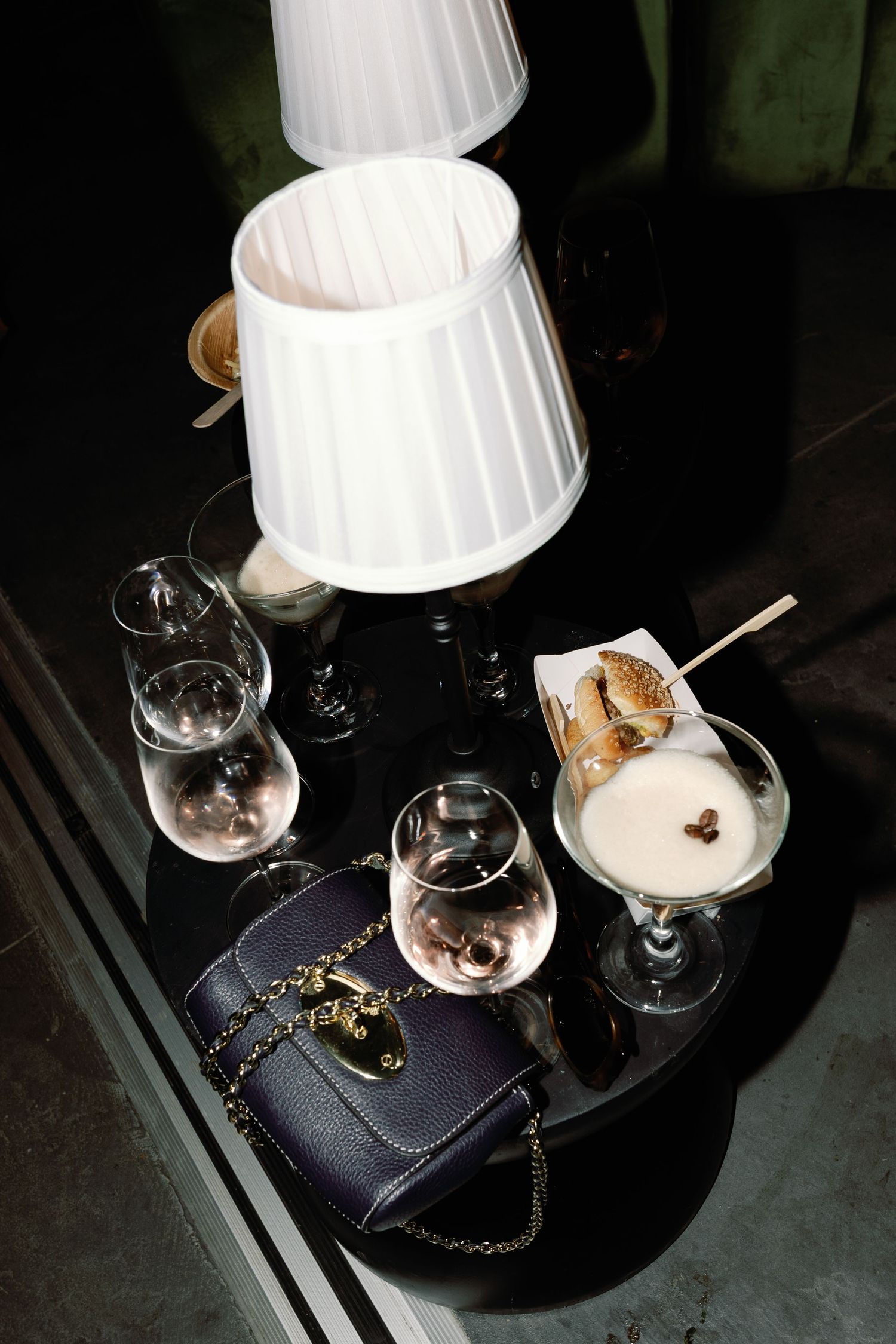 Dark romantic table setting with lamp, wine glasses, handbag, and creamy cocktail at wedding reception.
