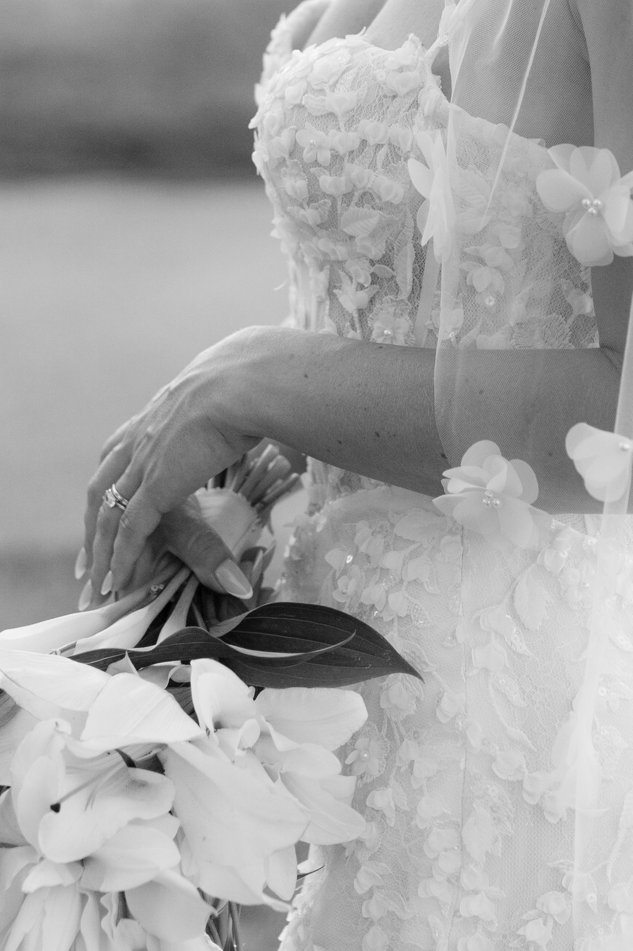 Black and white photo of a bride in a floral lace gown holding a wedding bouquet with a diamond ring visible.