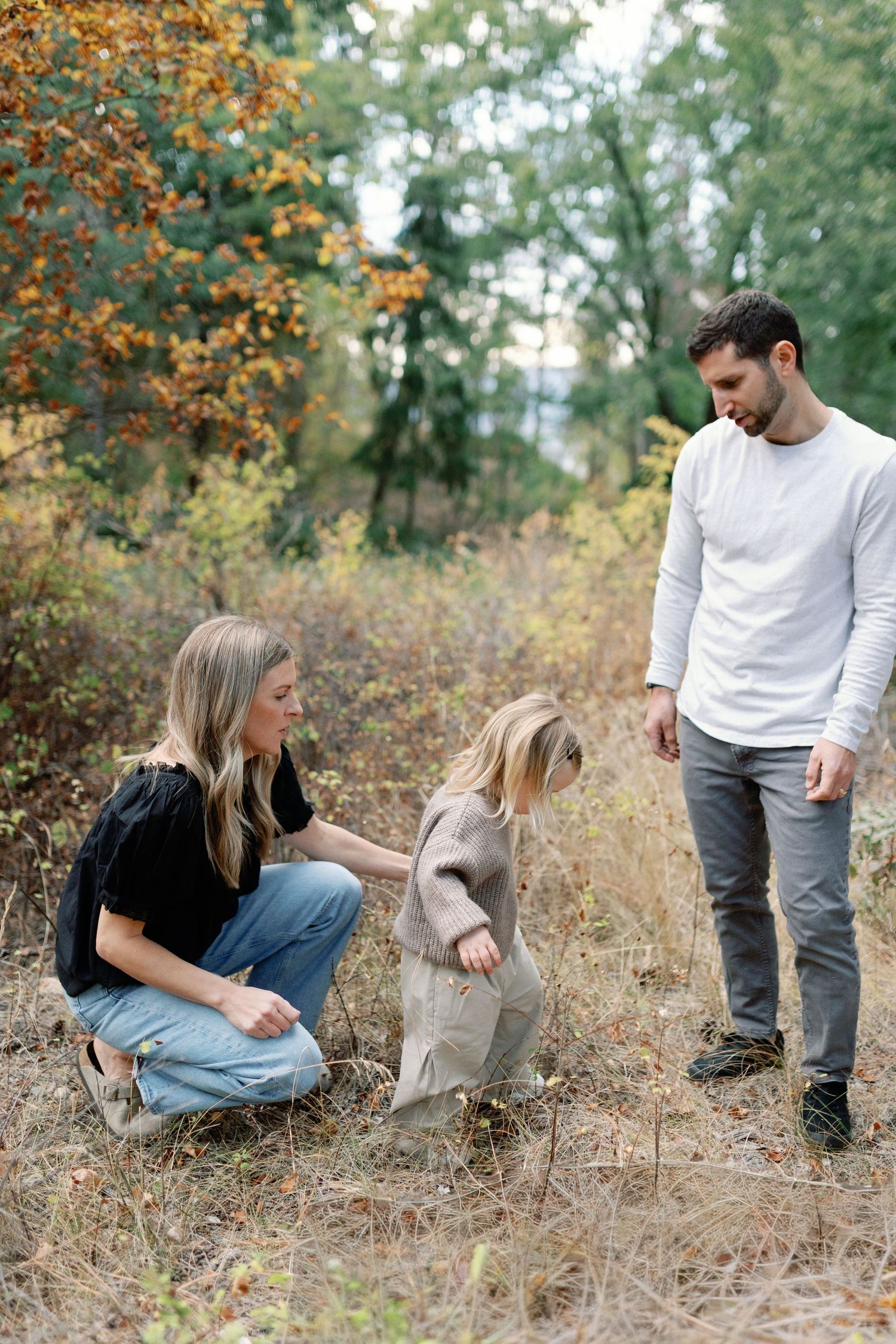 Family of three exploring autumn forest, parents watching young child walk through dry grass among colorful fall foliage.
