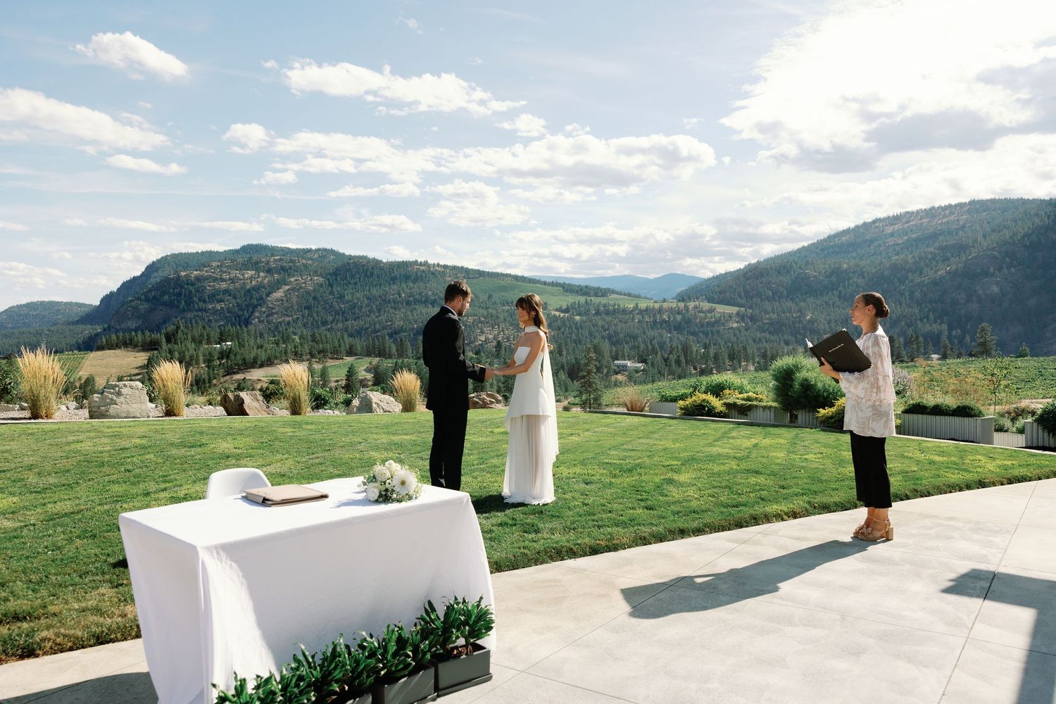 Intimate outdoor wedding ceremony on a patio with mountain landscape, officiant, bride, and groom holding hands.