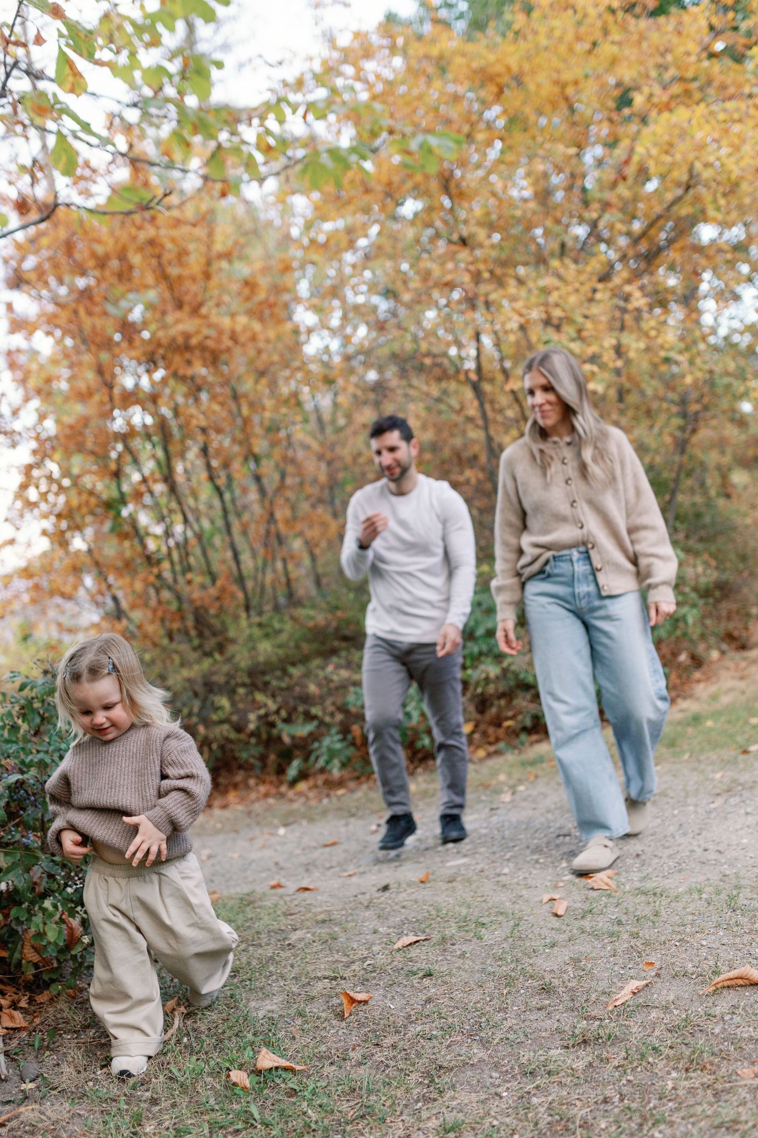 Family of three enjoying an autumn walk in a colorful fall forest, with young child exploring ahead of parents.
