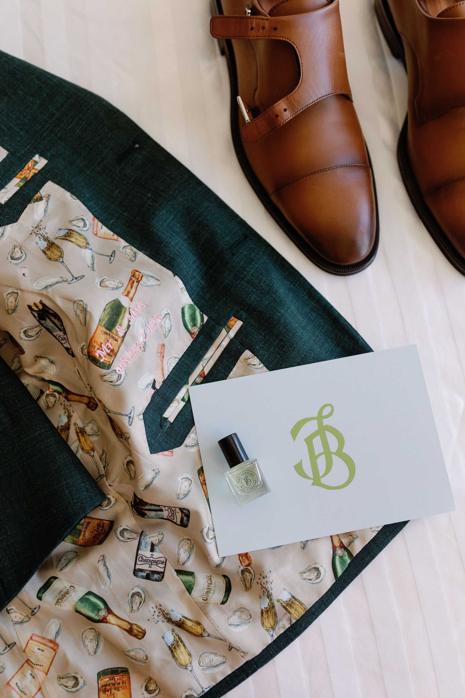Wedding flat lay featuring green suit, patterned lining, monogram card, cologne, and brown leather dress shoes.