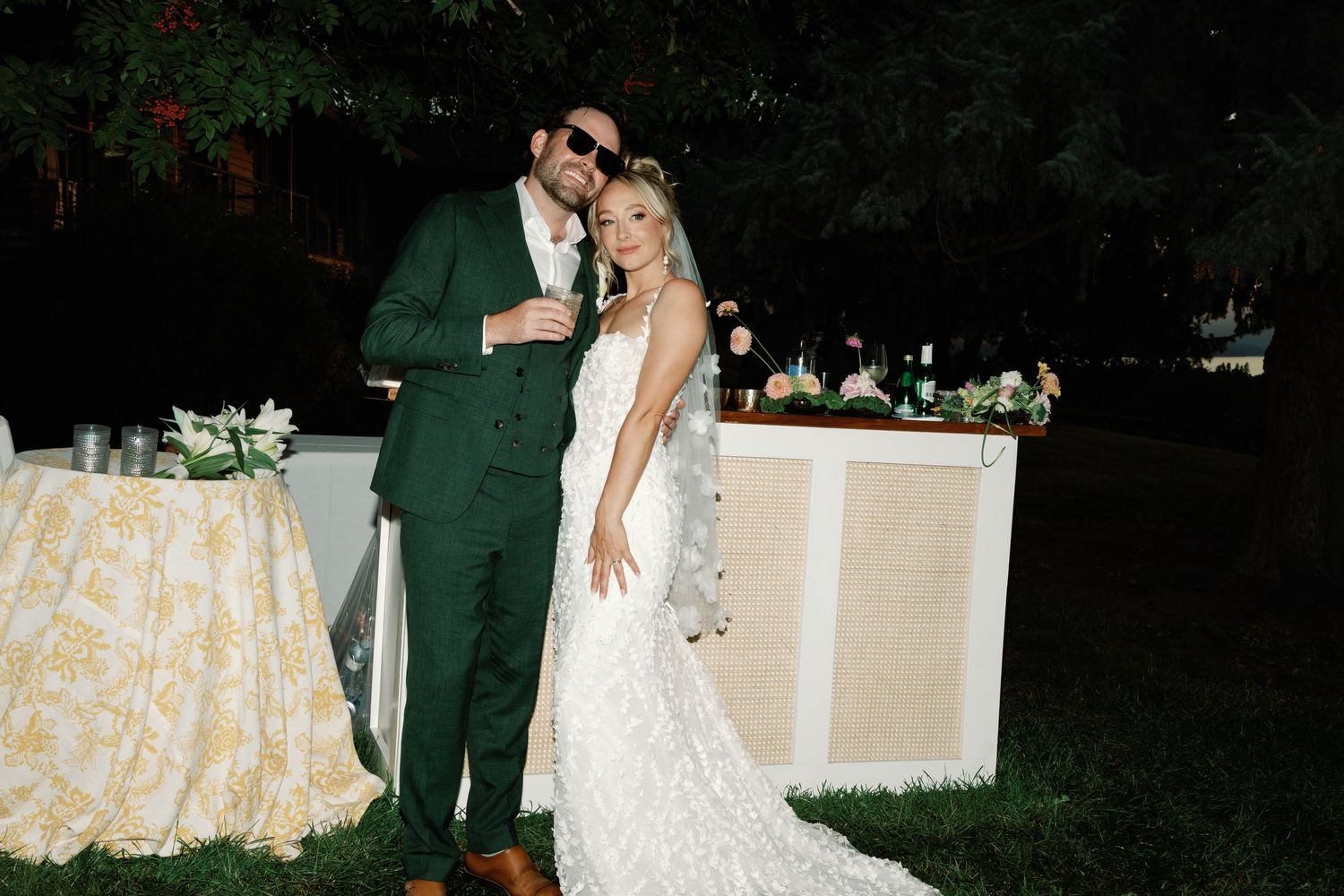 Bride and groom posing together outdoors at night, groom in green suit, bride in lace wedding gown.