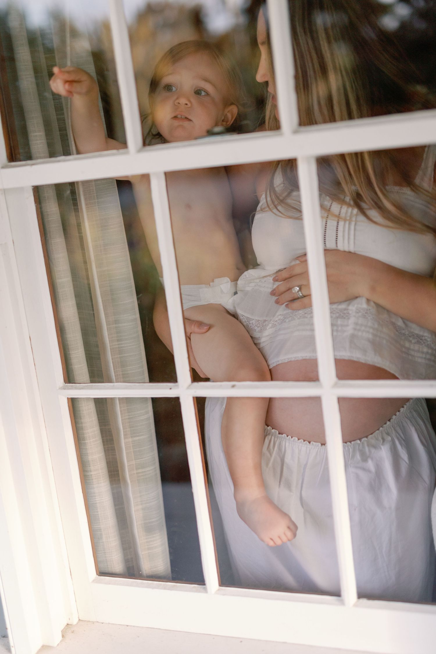 Pregnant woman holding toddler viewed through white-framed window from outside.
