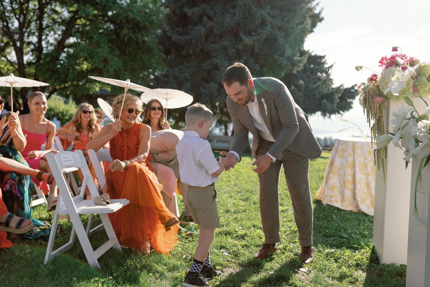 Groom in grey suit bending down to greet a young child during outdoor wedding ceremony.
