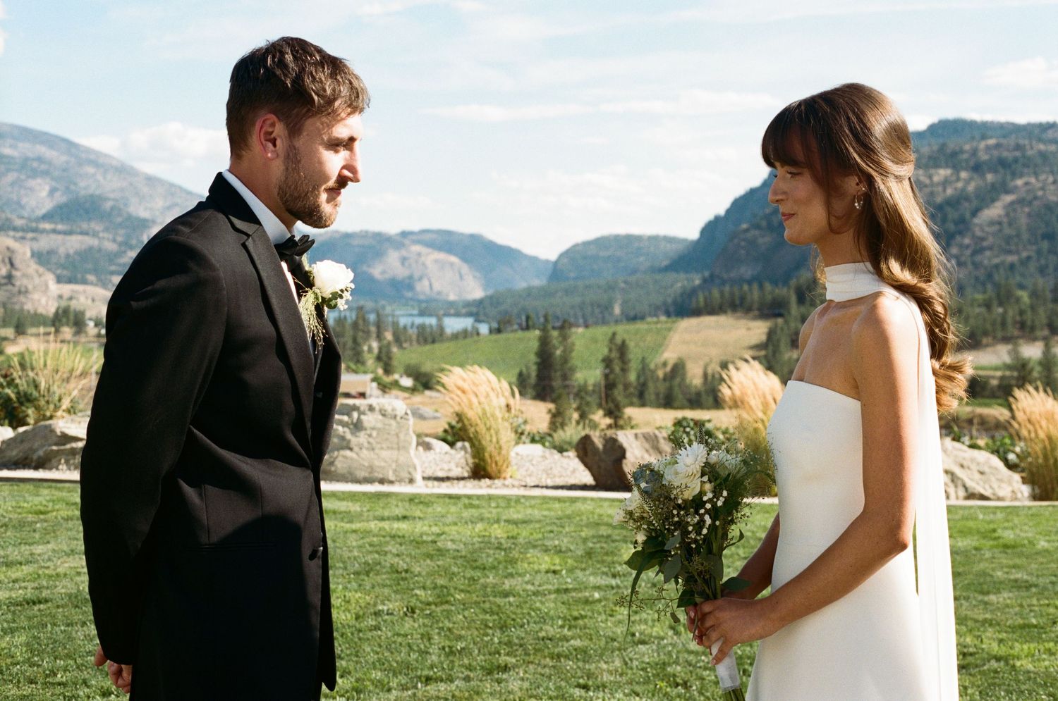 Smiling groom in black tuxedo faces bride holding white bouquet with mountain scenery behind them.