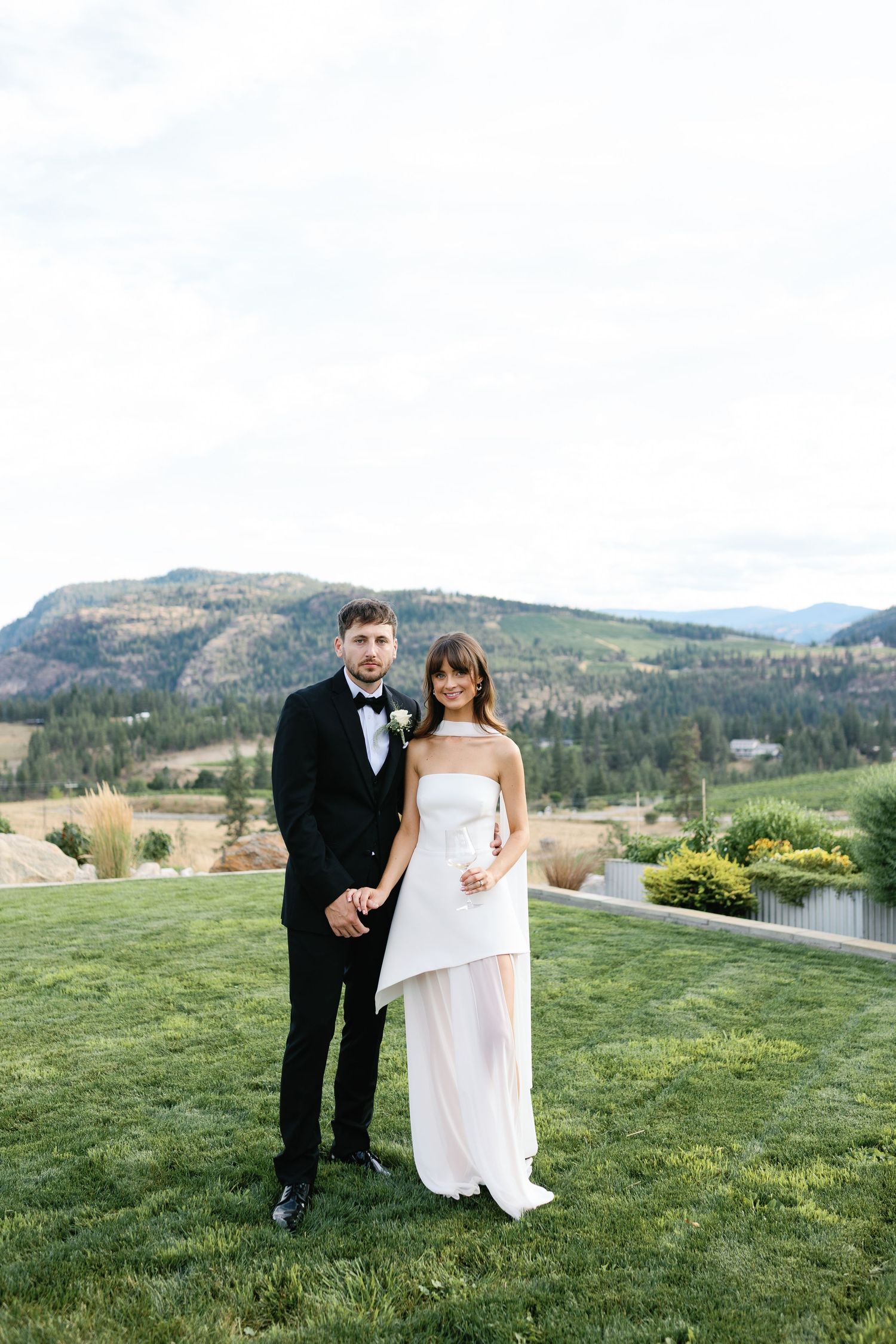 Bride and groom posing together on green lawn with stunning mountain valley backdrop.